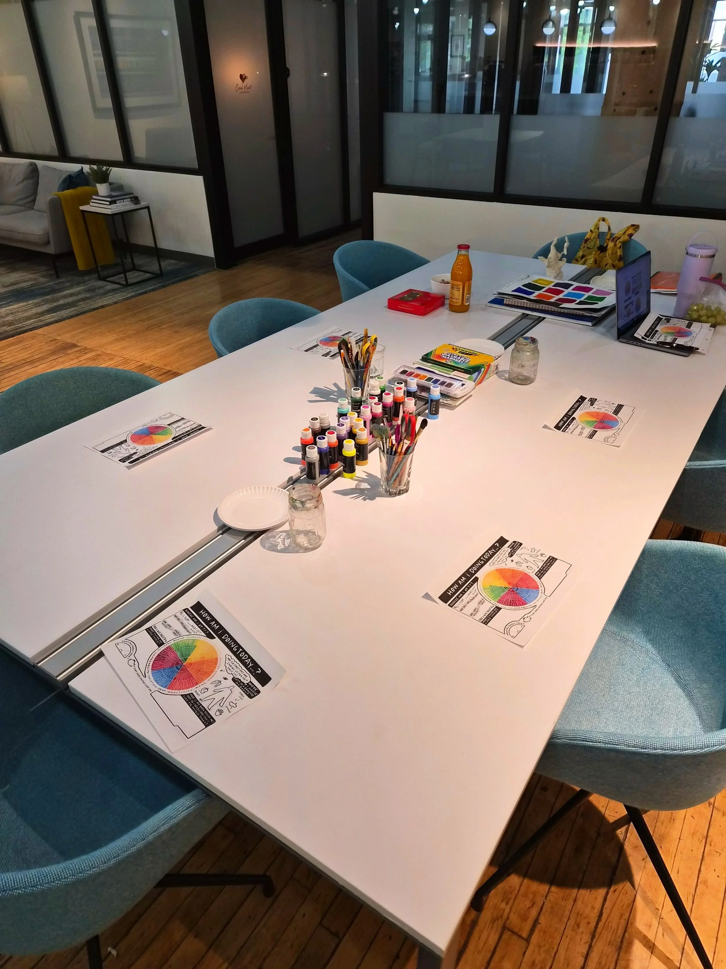 A white table with chairs, art supplies including colored markers, paint palettes, and color charts, in a room with glass walls and wooden floors.