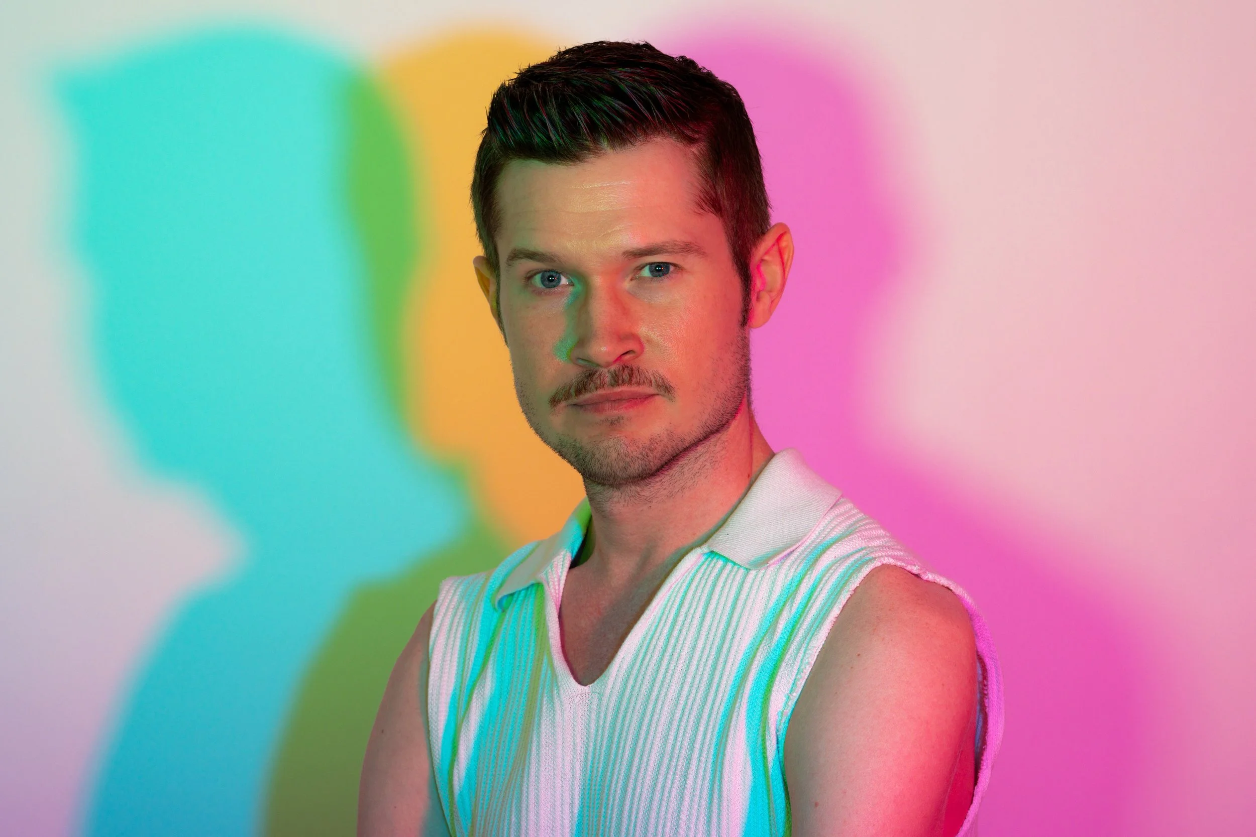 Editorial self-portrait of Christian S. Adams in a white sleeveless knit top, lit by overlapping cyan, yellow, green, and pink projections.