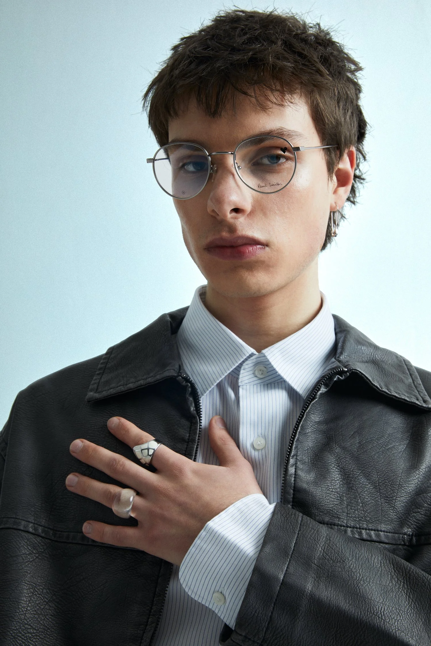 A young man wearing round glasses with a Paul Smith logo, a leather jacket, a white striped shirt, and multiple rings, standing against a plain light background.