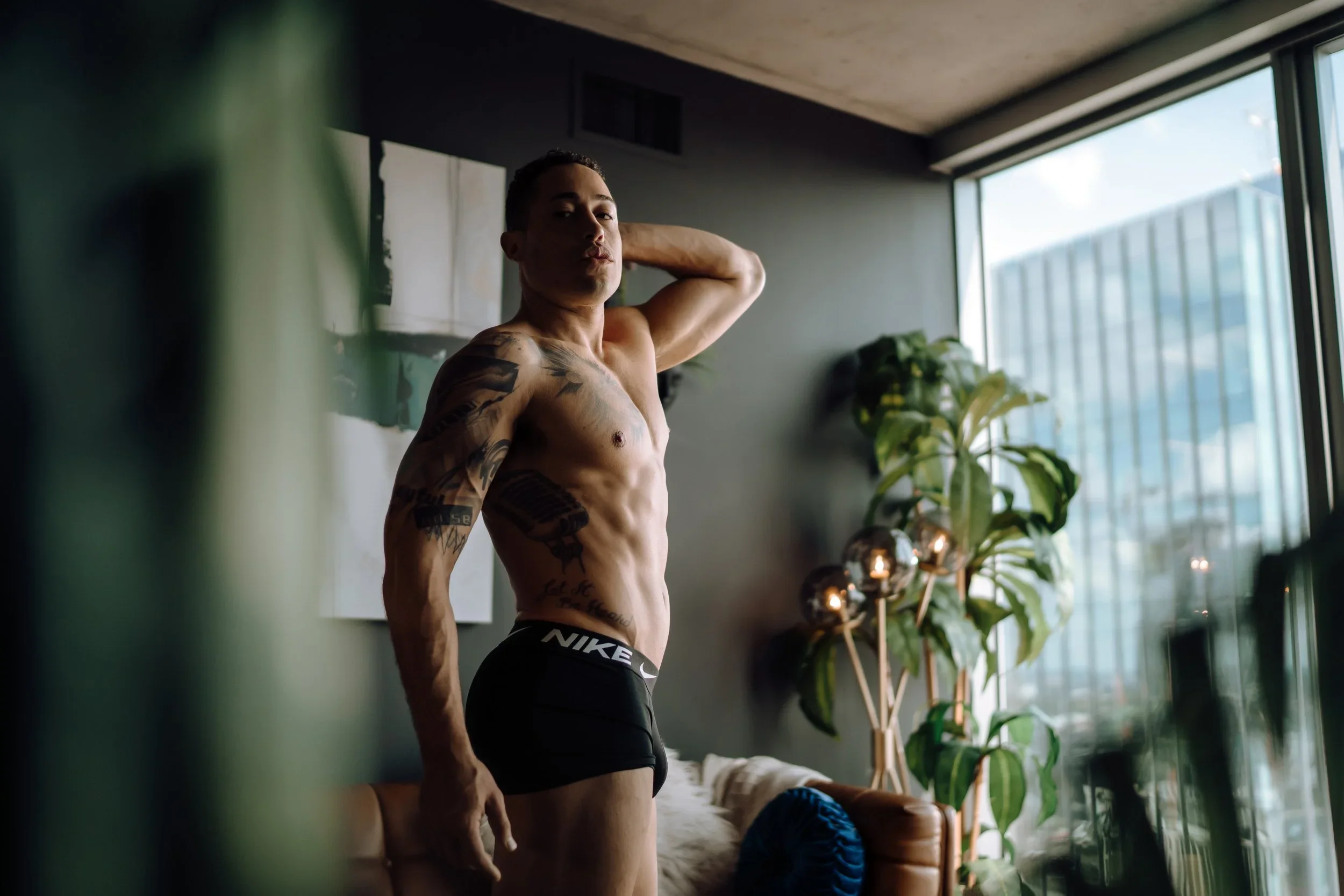 A shirtless man with tattoos stands in a modern, well-lit room with large windows, posing with one hand behind his head. There is a plant and some decorative lighting in the background.