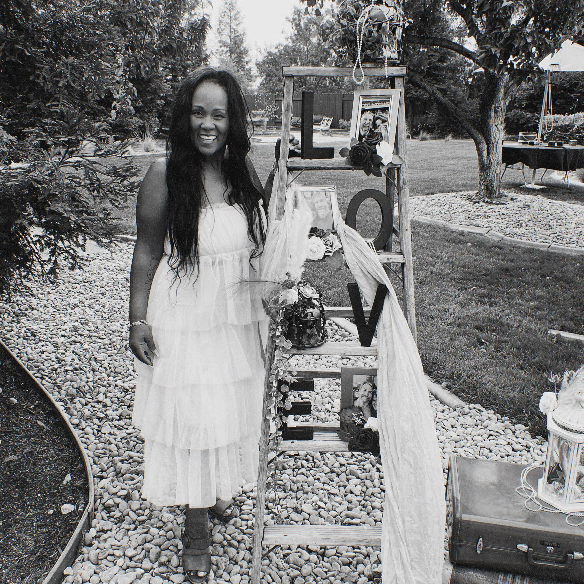 A smiling woman in a white dress standing in a garden, next to a wooden display with framed photos, roses, and decorative beads hanging from a frame, with trees, a table, and a pool in the background. Stacey runs The Palm House & Bohemian House