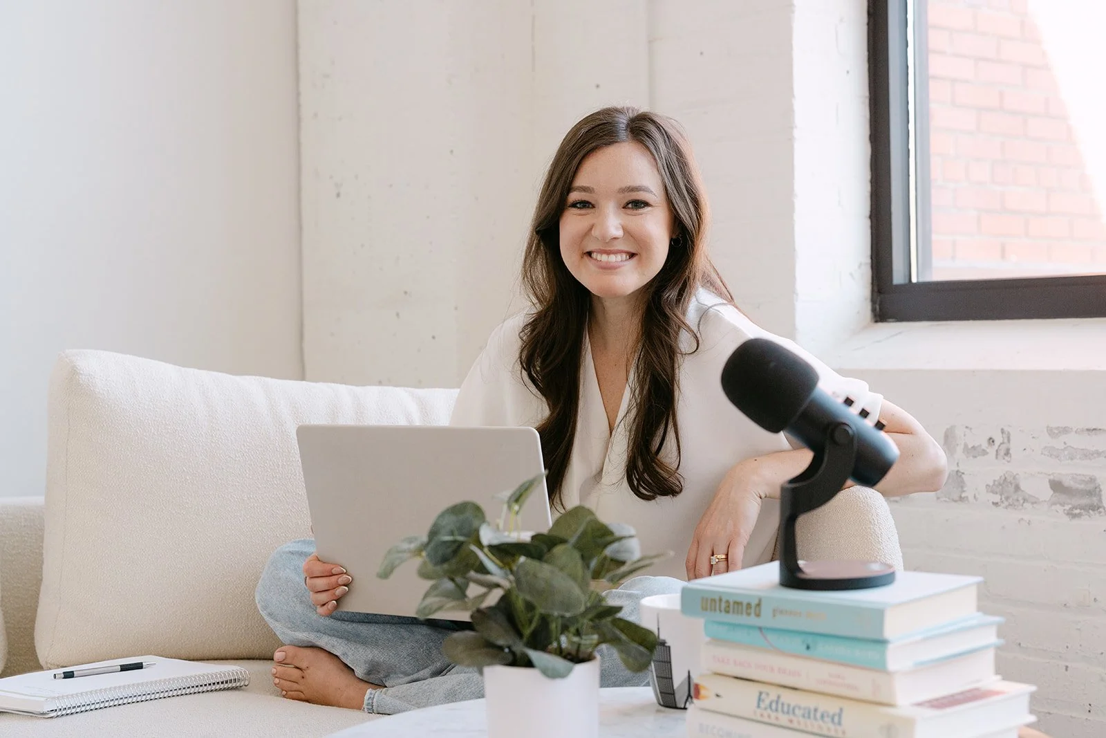 A woman with long brown hair sitting on a light-colored couch, smiling, holding a laptop, with a stack of books, a plant, and a microphone on a table nearby, near a window with a brick wall outside.