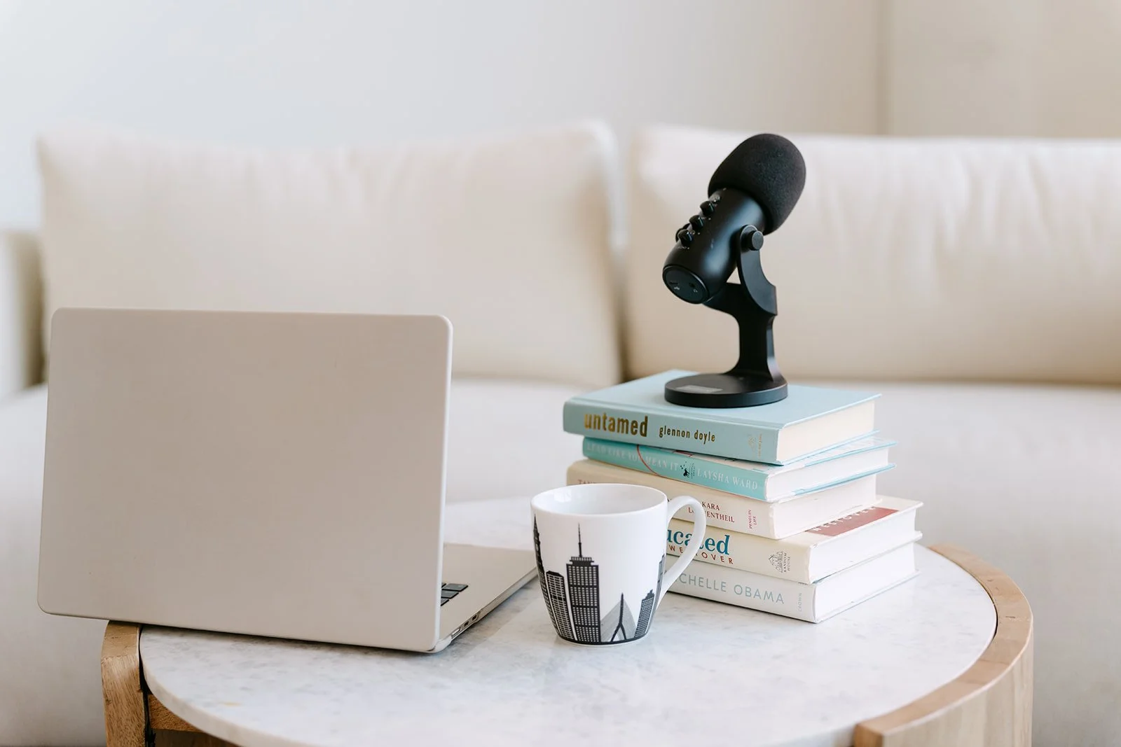 A white laptop, a white mug with a city skyline, a stack of books, a microphone, and a closed laptop on a round side table in a bright, neutral-colored room.