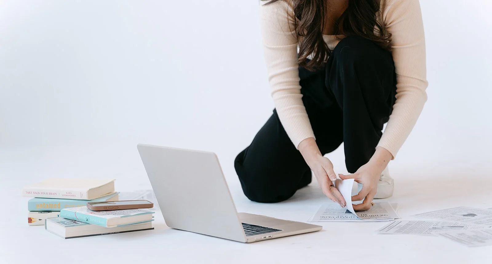 Person crouched on the floor flipping through papers, surrounded by books, a closed laptop, and a smartphone on a white surface.