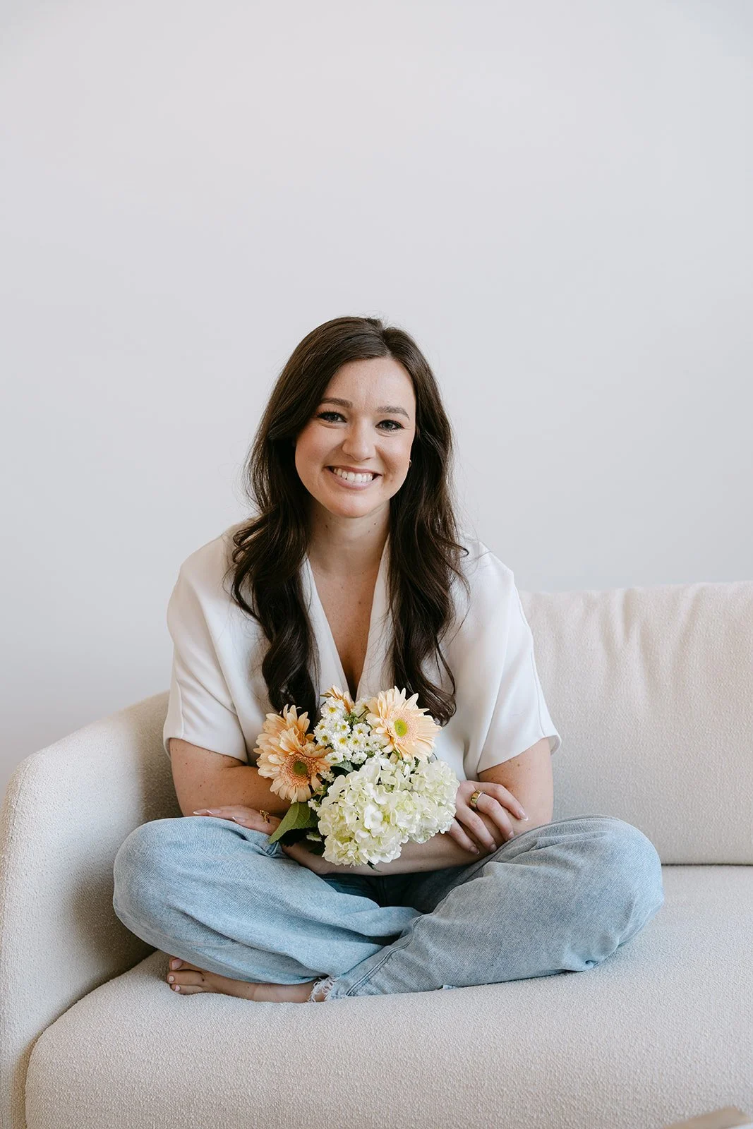 A woman sitting cross-legged on a beige couch, holding a bouquet of flowers, smiling at the camera in a light-colored room.