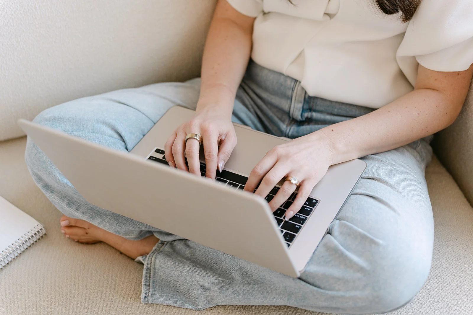 Person sitting cross-legged on a beige couch using a silver laptop, wearing a white shirt and light blue jeans.