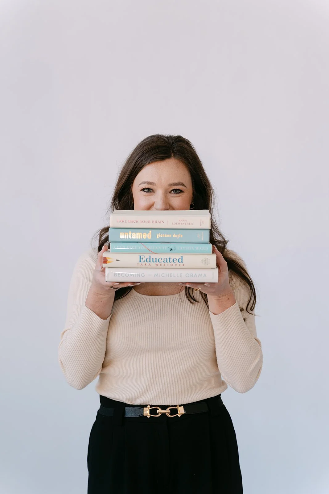 A woman standing against a plain light background, holding a stack of books in front of her face, smiling with only her eyes visible.