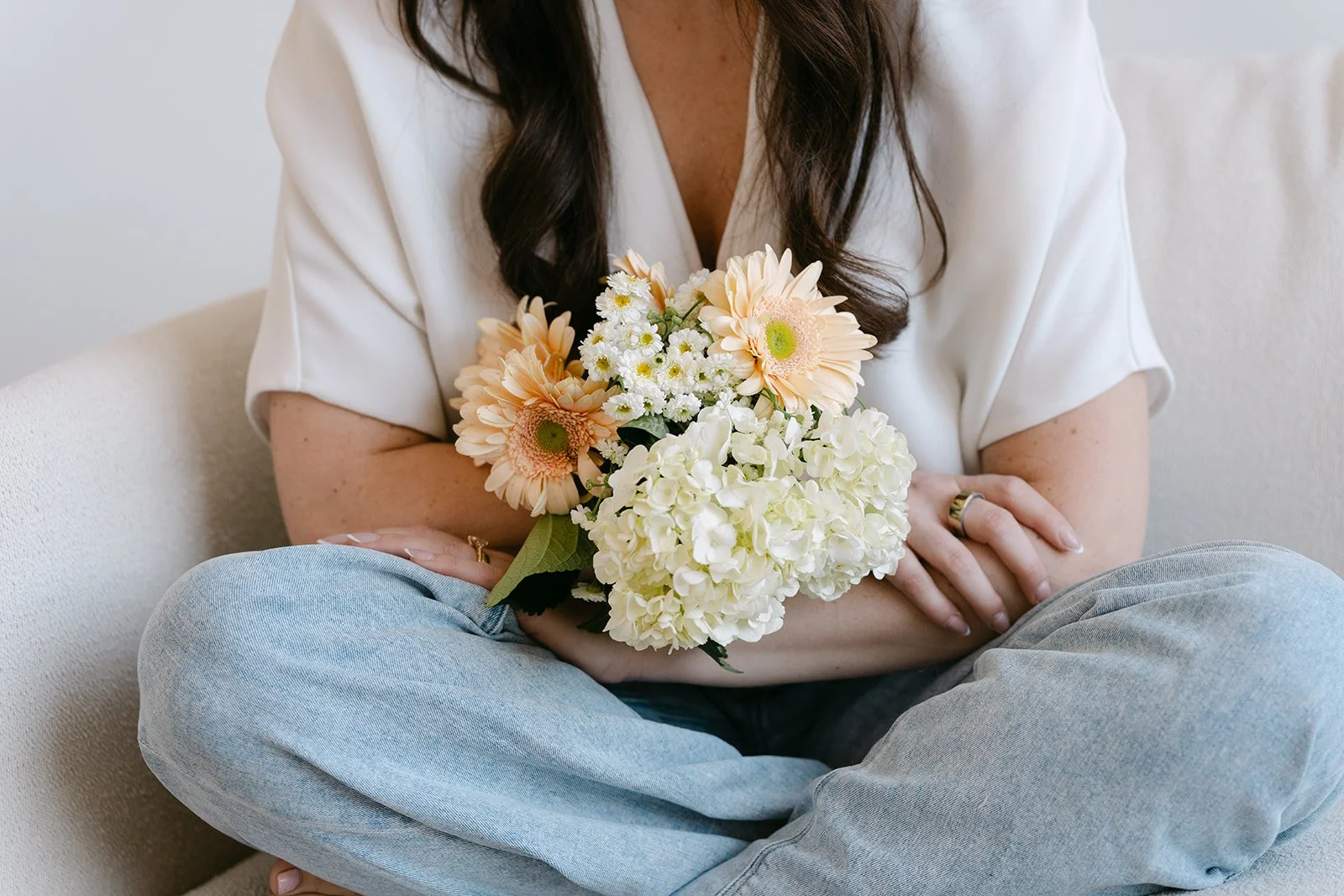 A woman with long dark hair, wearing a white top and light blue jeans, sitting cross-legged on a beige sofa, holding a bouquet of cream, white, and pale peach flowers, including hydrangeas and daisies.