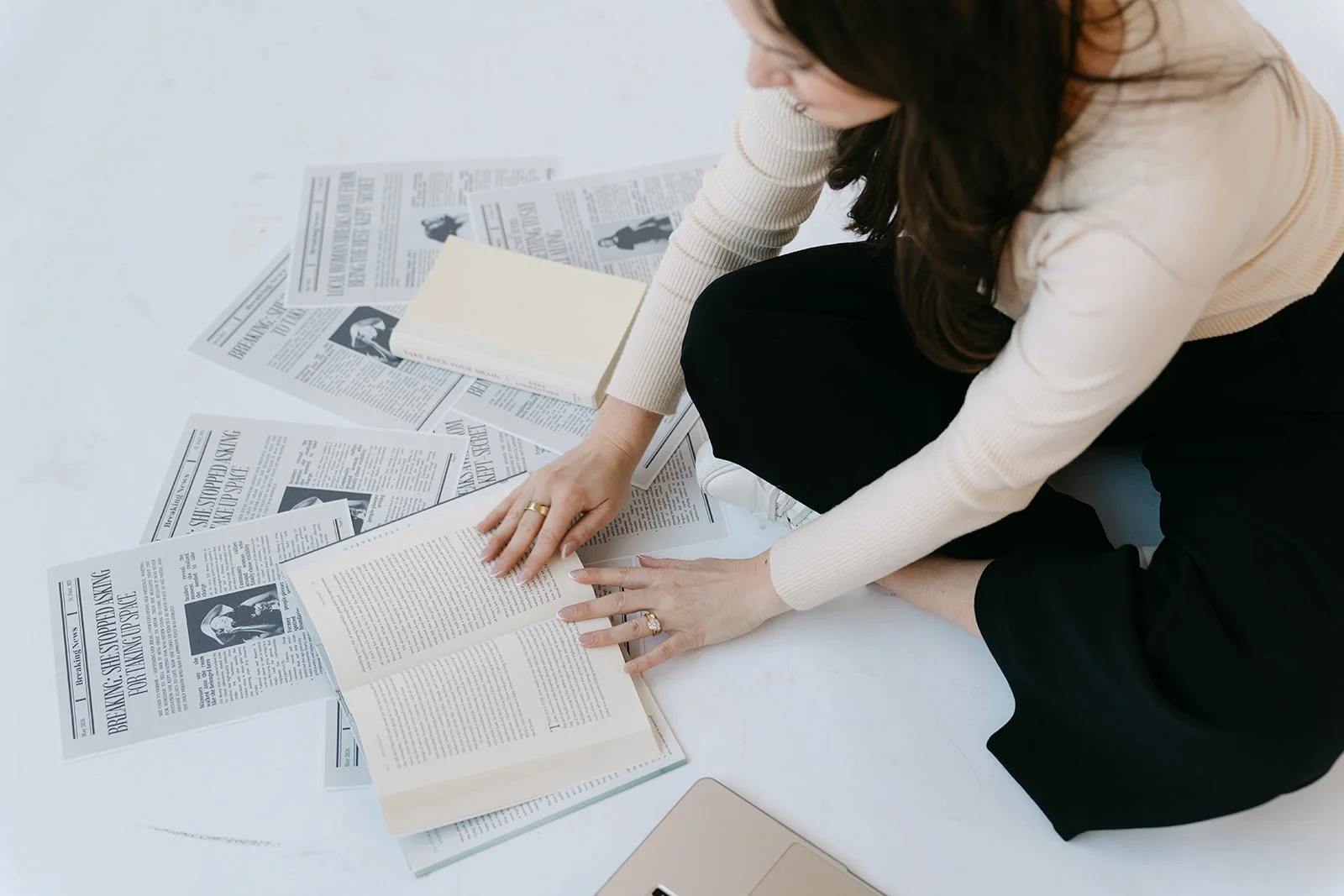 A woman sitting on the floor, reading a book, surrounded by scattered newspapers.