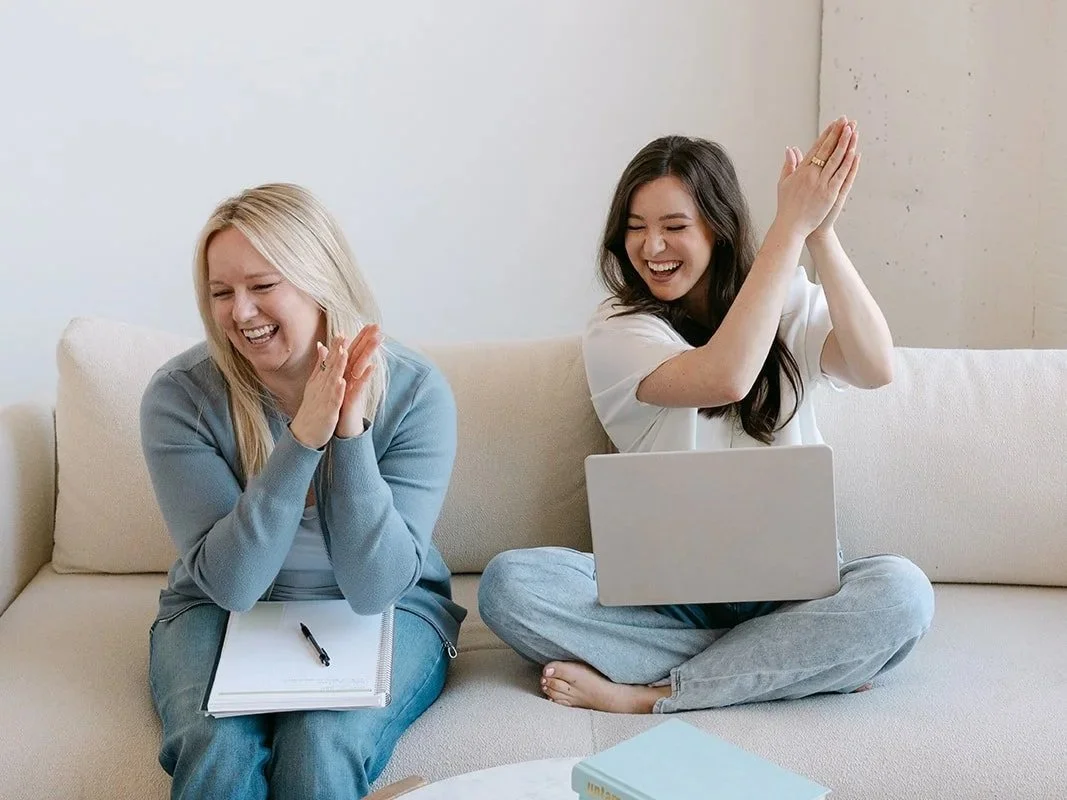 Two women sitting on a beige couch, laughing and clapping, one with a notepad and pen, the other with a laptop, in a casual indoor setting.