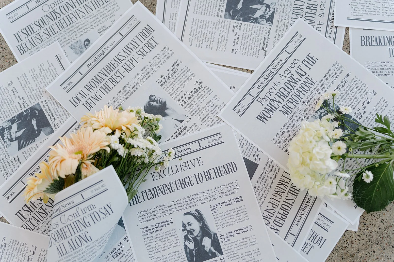 Several scattered black-and-white newspapers on a surface with two small bouquets of white and peach flowers.