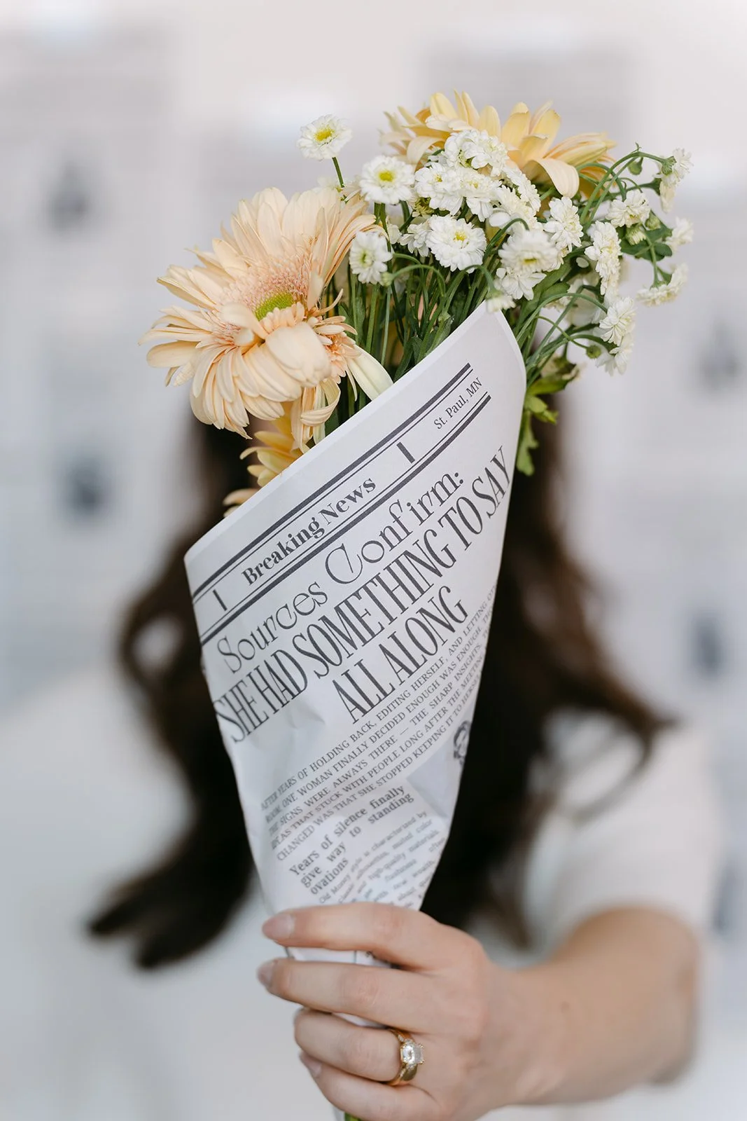 A person holding a bouquet of white and peach flowers wrapped in a newspaper with headlines about a firm and legal issues.