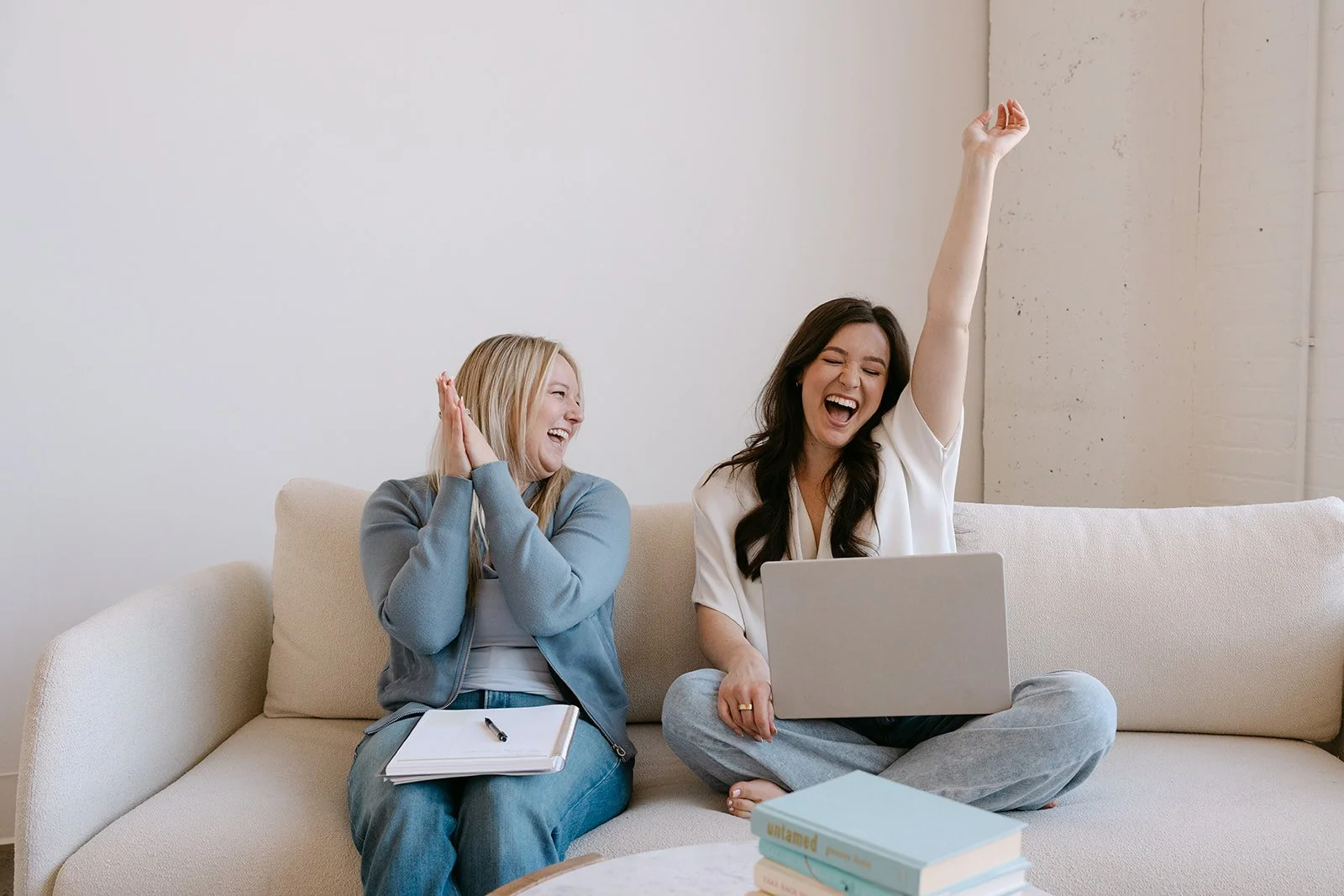 Two women sitting on a beige couch, laughing and celebrating while looking at a laptop. One woman, with dark hair, is raising her hand in excitement, while the other woman, with blonde hair, claps and smiles.