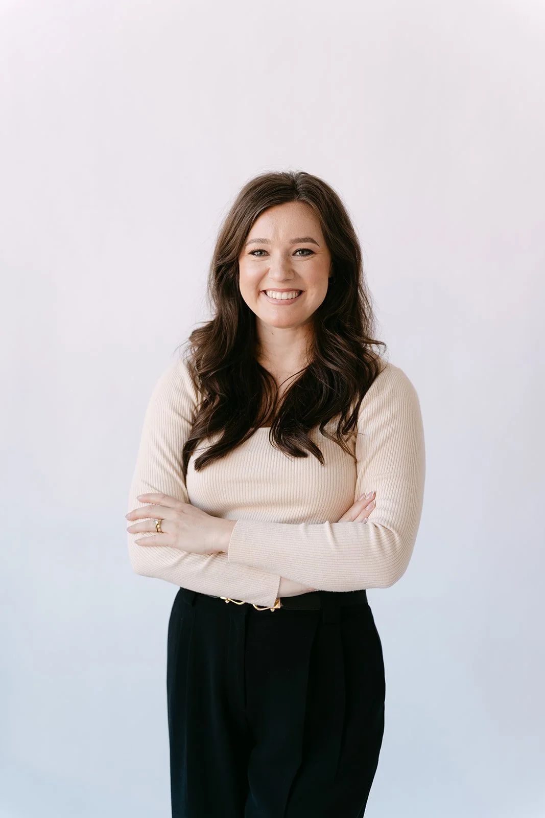 A woman with long dark hair smiling with arms crossed, wearing a beige ribbed long-sleeve top and black pants, standing in front of a plain light background.