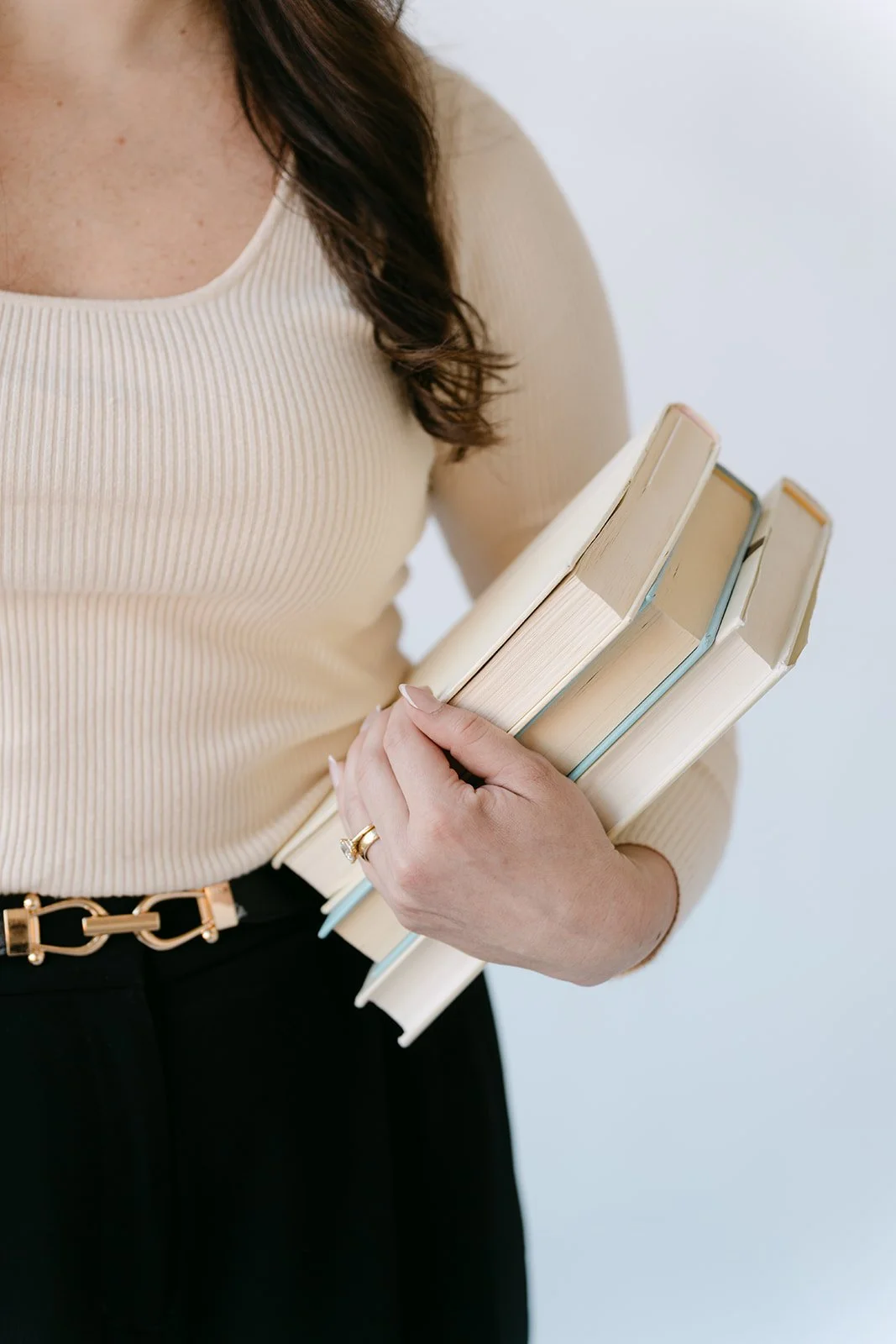 A woman holding three beige books with one hand, wearing a cream-colored ribbed top and a black skirt with a gold belt.