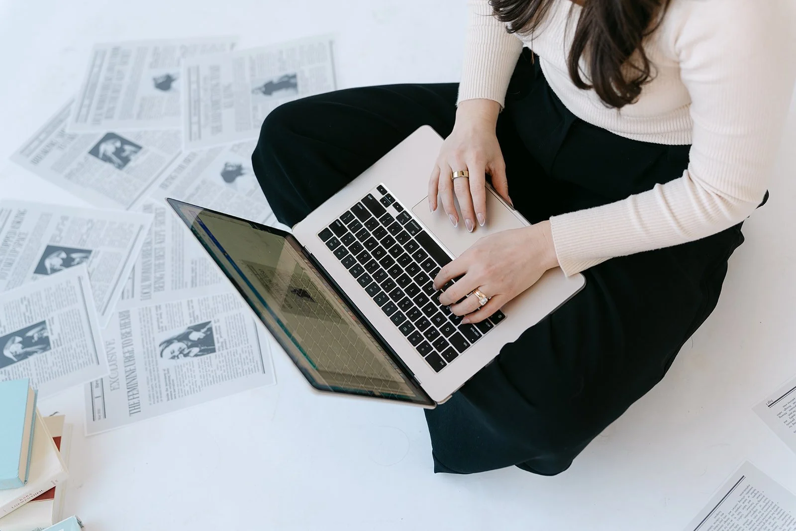Person sitting at a white table with scattered newspapers and working on a silver laptop.