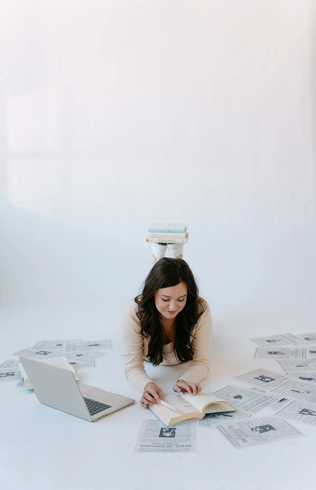 Woman lying on the floor reading a book surrounded by scattered newspapers, with a laptop open beside her