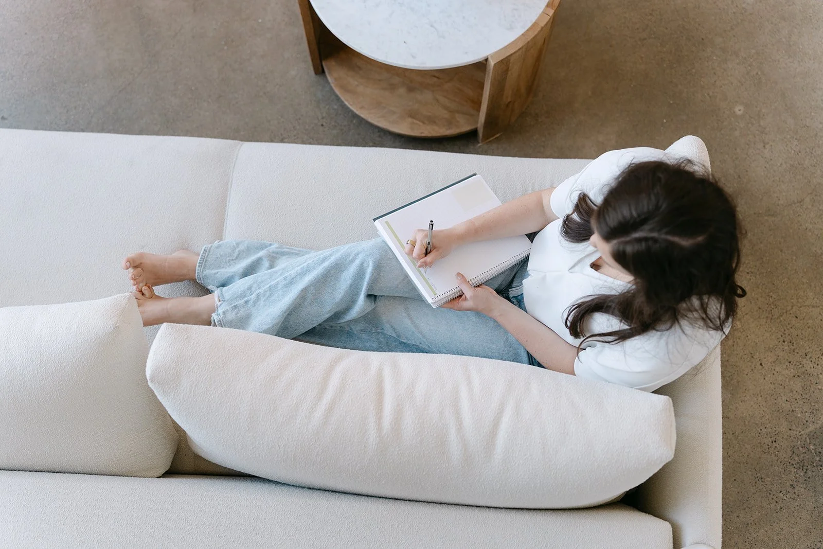 Top view of a woman sitting on a beige couch, writing in a notebook with a pen. She is wearing a white shirt and light blue jeans. A round wooden coffee table is in the background, and her legs are crossed, with her bare feet resting on the couch.