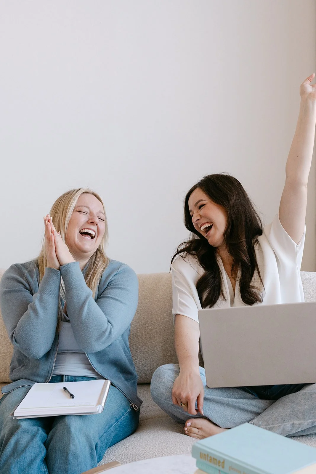 Two women sitting on a beige couch, laughing and enjoying each other's company; one with blonde hair in a blue jacket and notebook, the other with dark hair raising her hand, using a laptop.