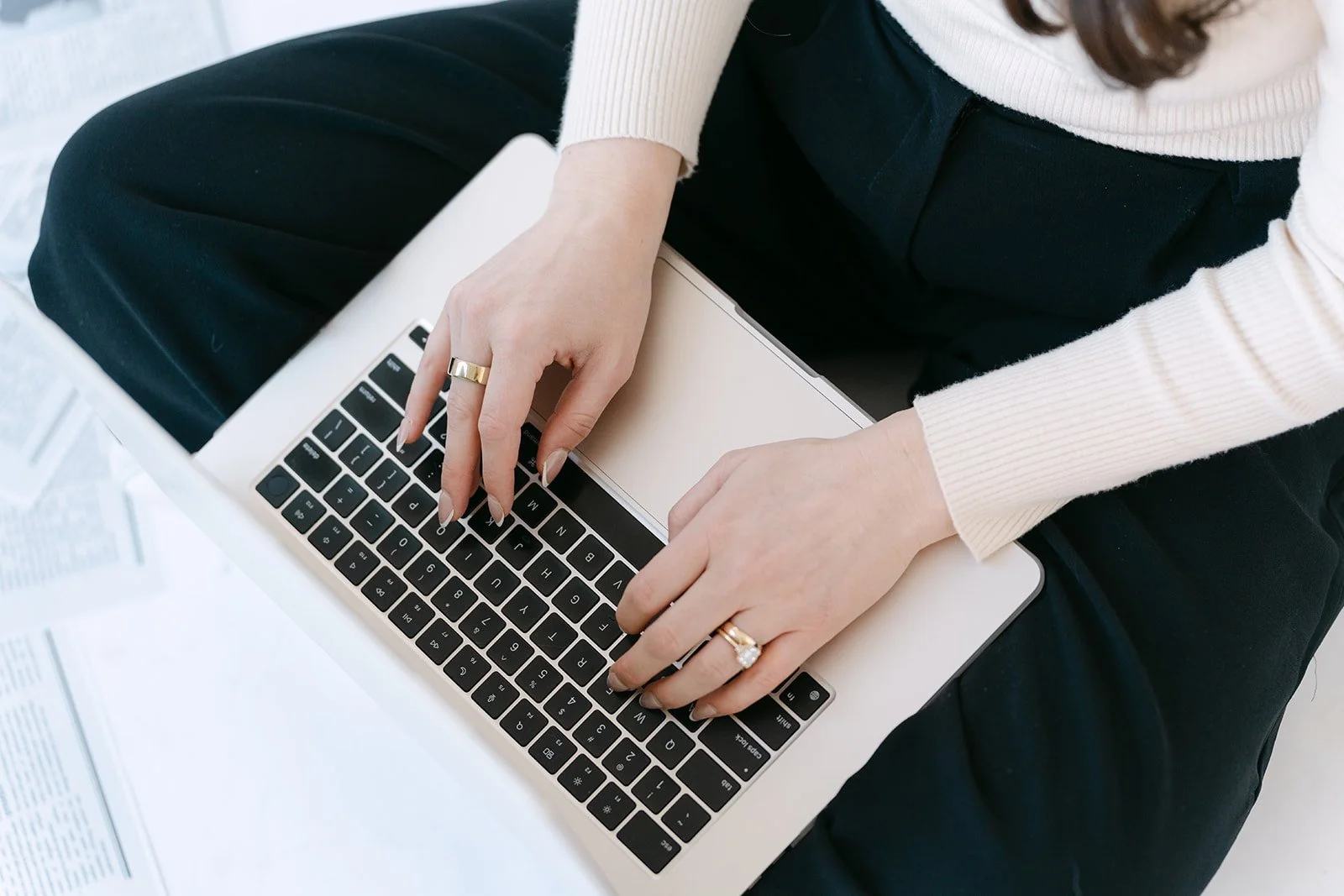 A person wearing a light-colored sweater and multiple rings on their fingers is sitting cross-legged and typing on a laptop keyboard. There are papers with printed text scattered nearby.