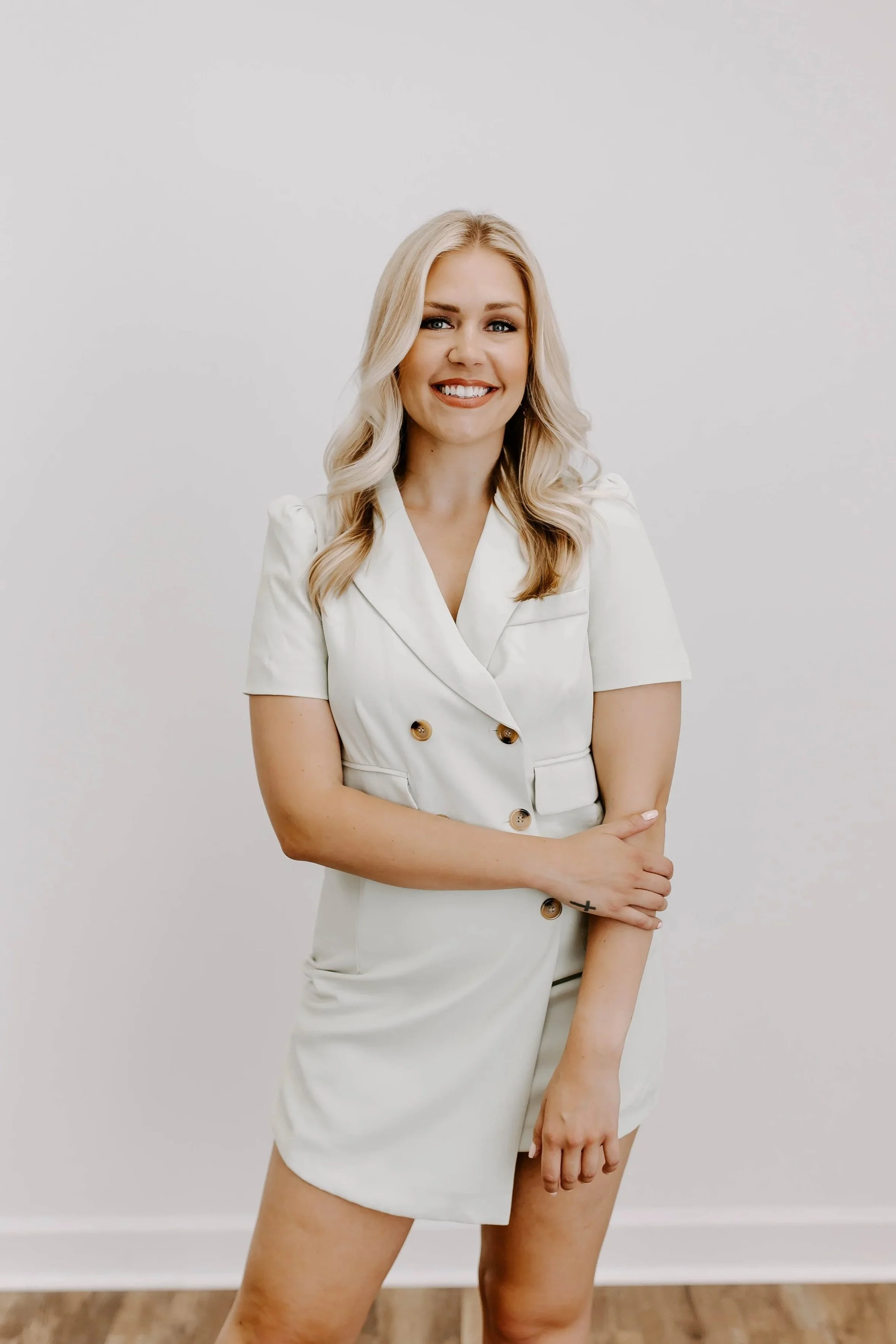 A woman with blonde hair, blue eyes, and light skin wearing a white short-sleeved dress with gold buttons, standing against a plain white background. She is smiling, with her arms crossed, and a small tattoo of a cross is visible on her left forearm.