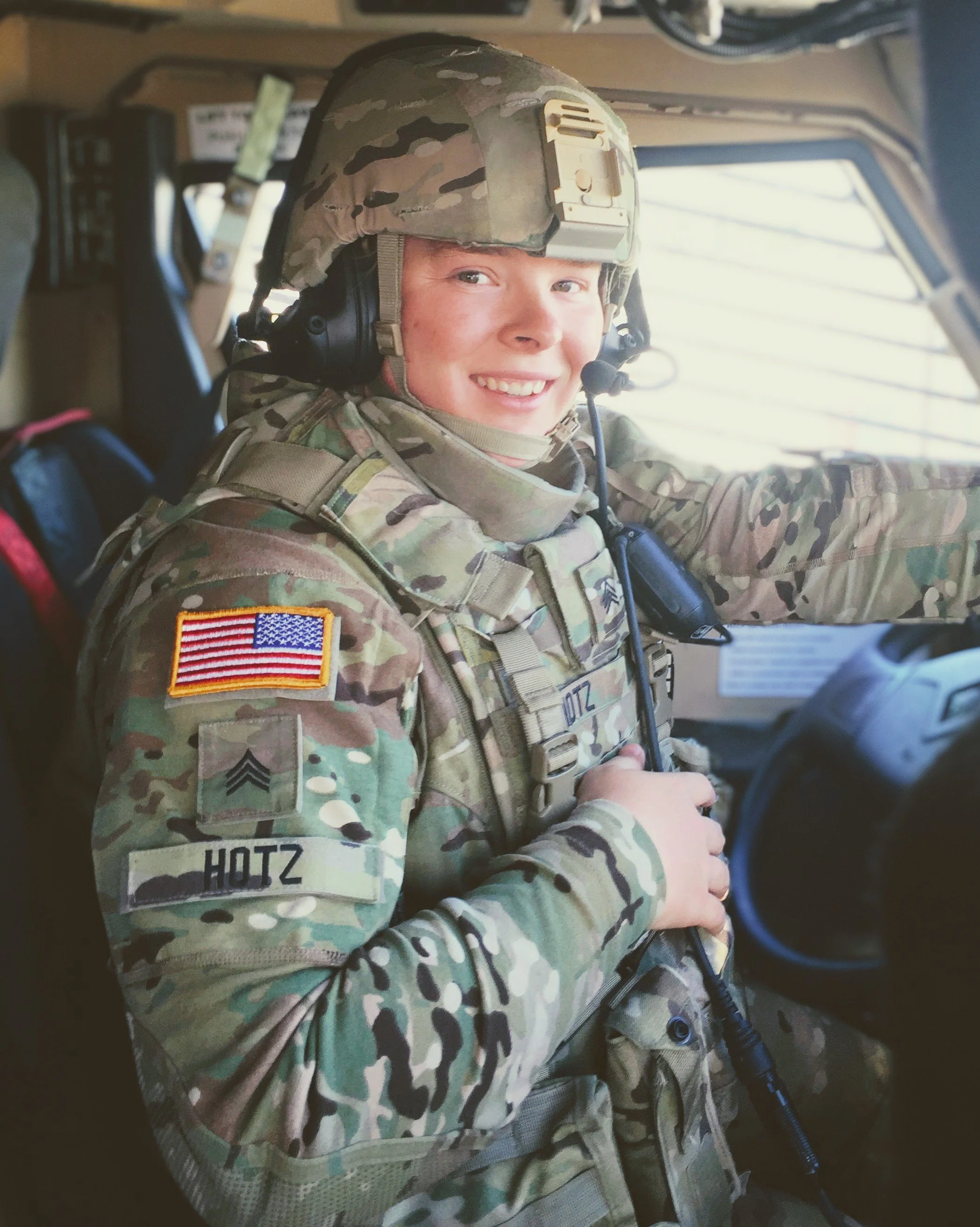 A smiling military soldier wearing a camouflage uniform, helmet, and headset, sitting inside a military vehicle.