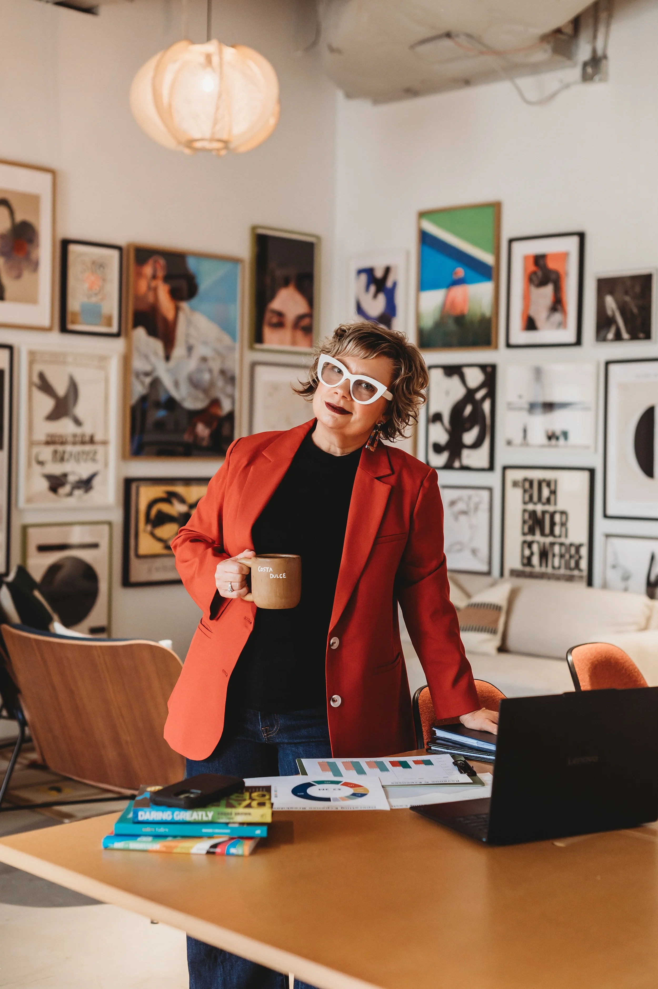 Woman wearing white glasses and a red blazer standing in an art gallery, holding a coffee mug, surrounded by framed artwork on the wall.