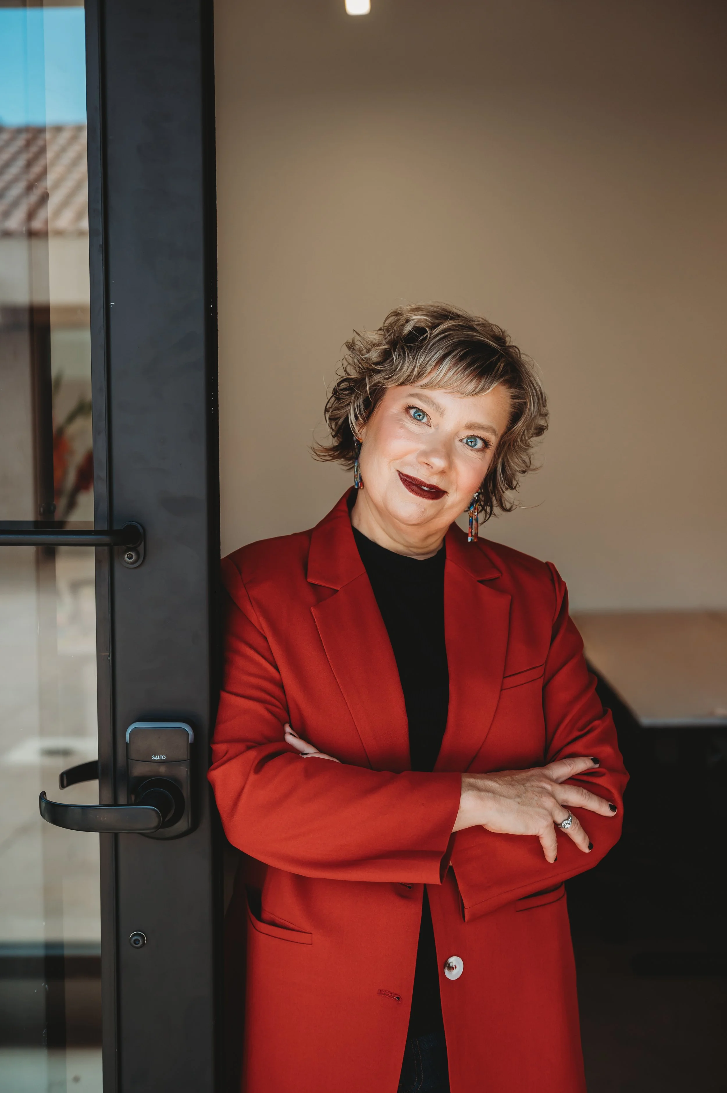 A woman with short, curly hair, wearing a red blazer and black top, standing with arms crossed and smiling at the camera.