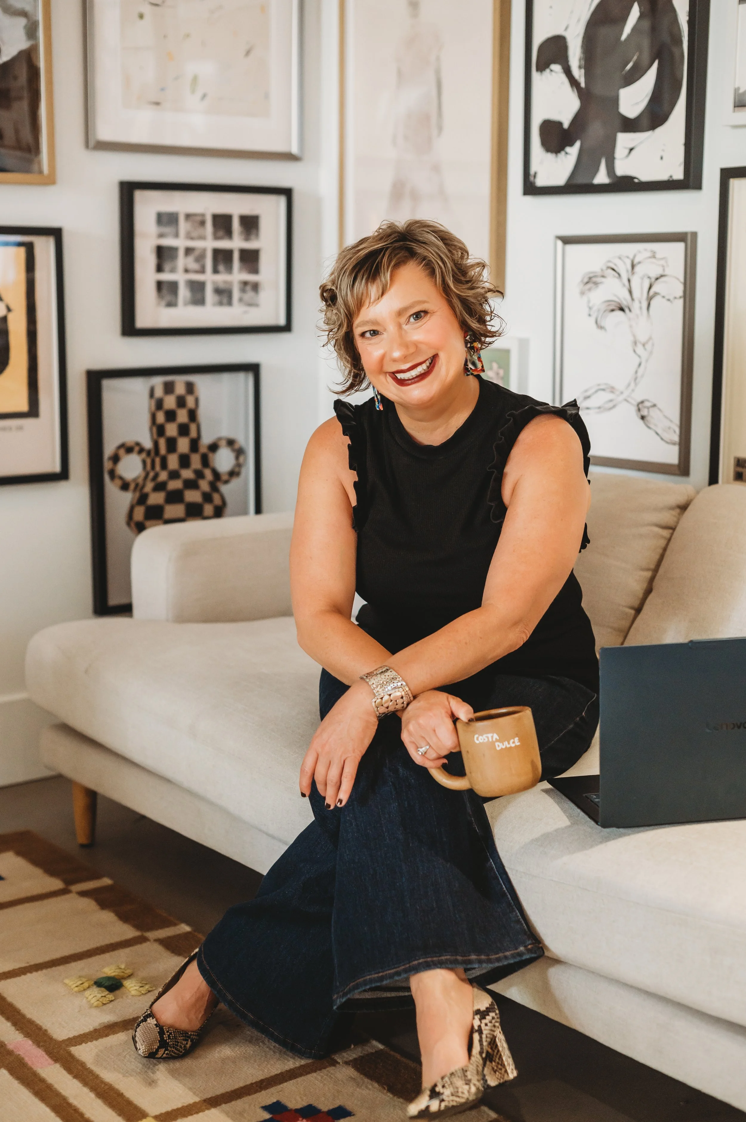 A woman with short curly hair is sitting on a beige sofa in an art gallery or living room. She is smiling and holding a coffee mug that has the words 'COSTA DULCE' written on it. She is wearing a sleeveless black top, a silver bracelet, and high heels with a snakeskin pattern. There are several framed artworks on the wall behind her, including a checkered pattern and abstract designs. The floor has a patterned rug, and there's a laptop on her lap.