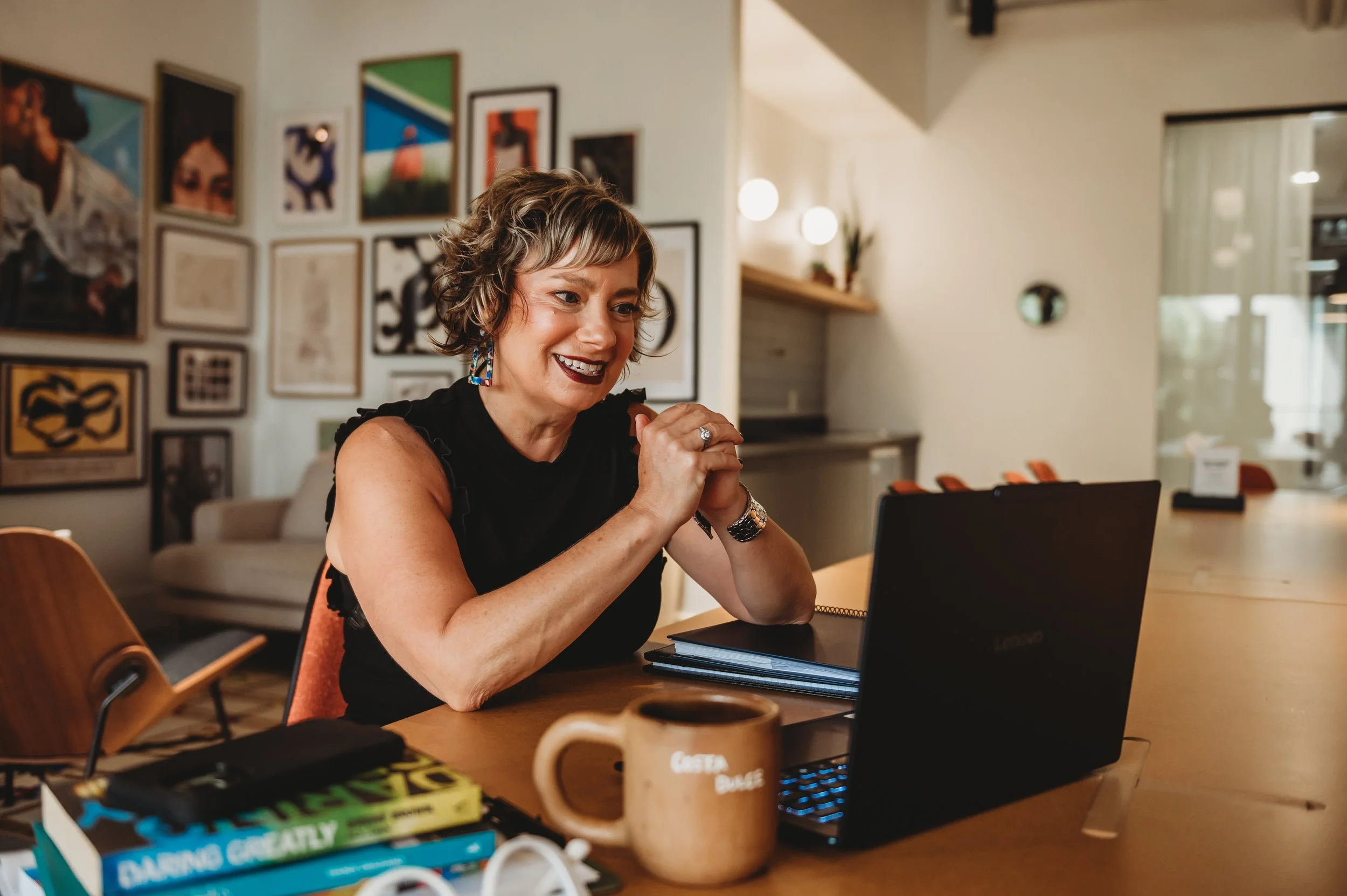 A woman with short wavy hair and colorful earrings, smiling and sitting at a table with a laptop and books in a cozy, well-lit room with artwork on the wall.