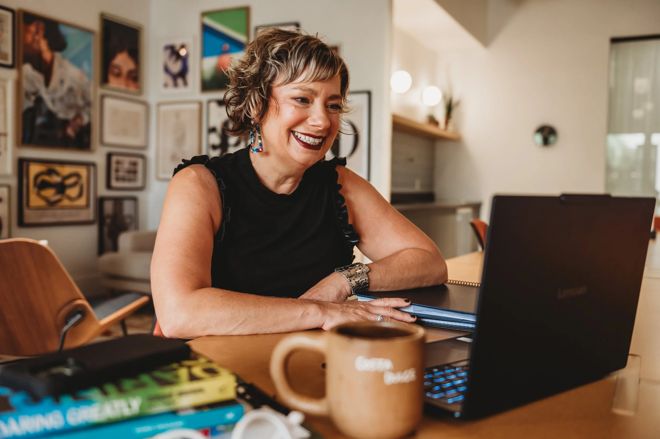 A woman with short curly hair and colorful earrings smiling while working on a laptop at a wooden table in a room decorated with framed artwork on the wall.