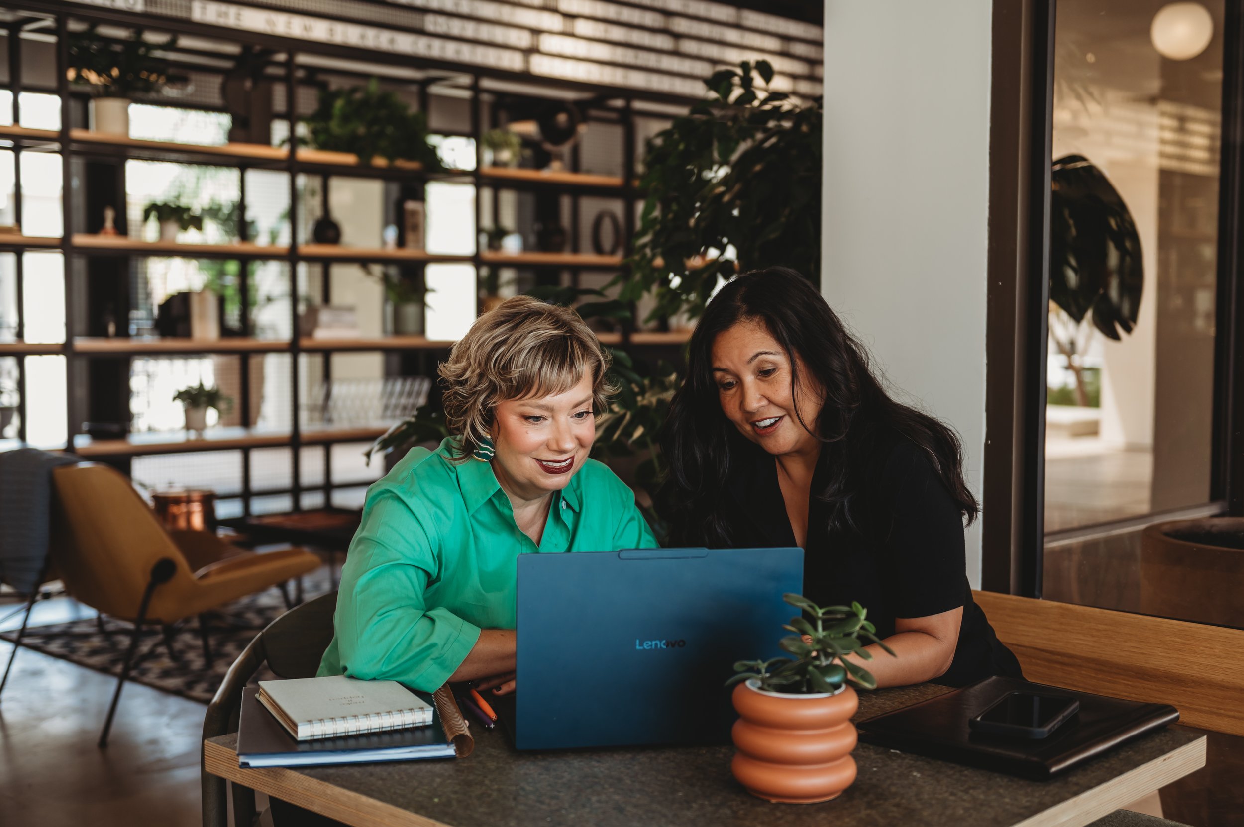 Two women sitting together at a table in a modern, well-lit cafe, looking at a laptop screen, with notebooks and a small potted plant on the table.