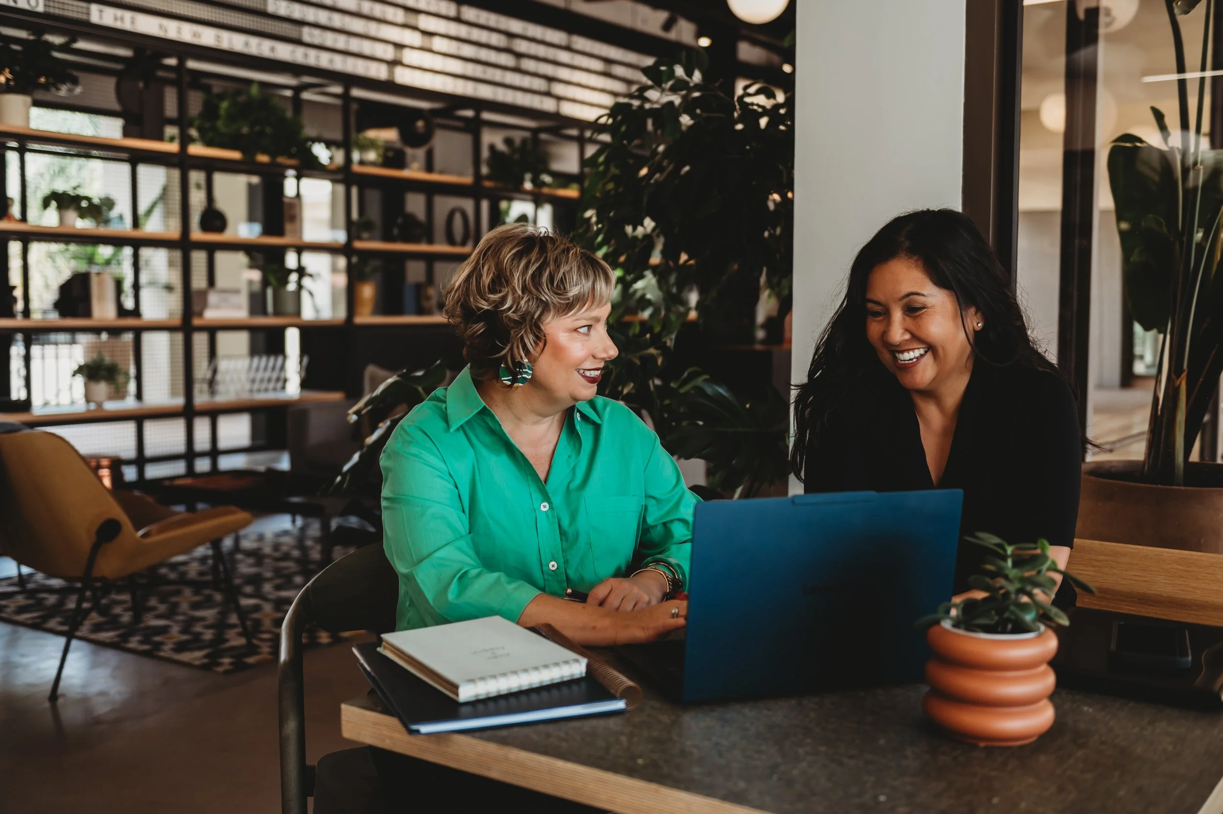 Two women are sitting at a table in a modern, well-lit cafe. They are smiling and looking at a laptop. The woman on the left has short, curly blonde hair and is wearing a green shirt. The woman on the right has long black hair and is wearing a black top. The table has notebooks and a potted plant. In the background, there are shelves with decorative items and large windows.