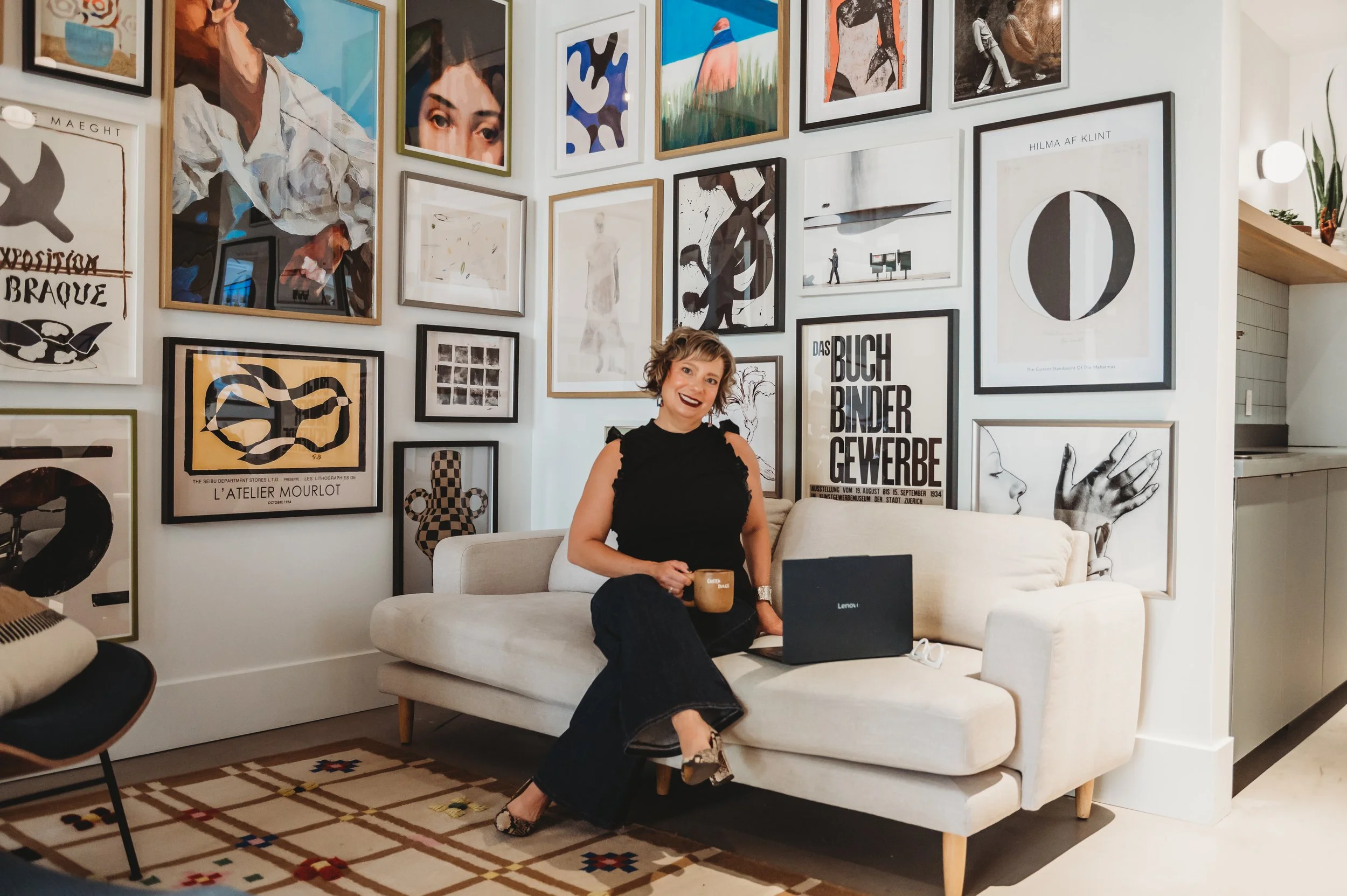 Woman with short, curly hair sitting on a white sofa with a latte in her hand in a living room decorated with numerous framed art prints on the wall.