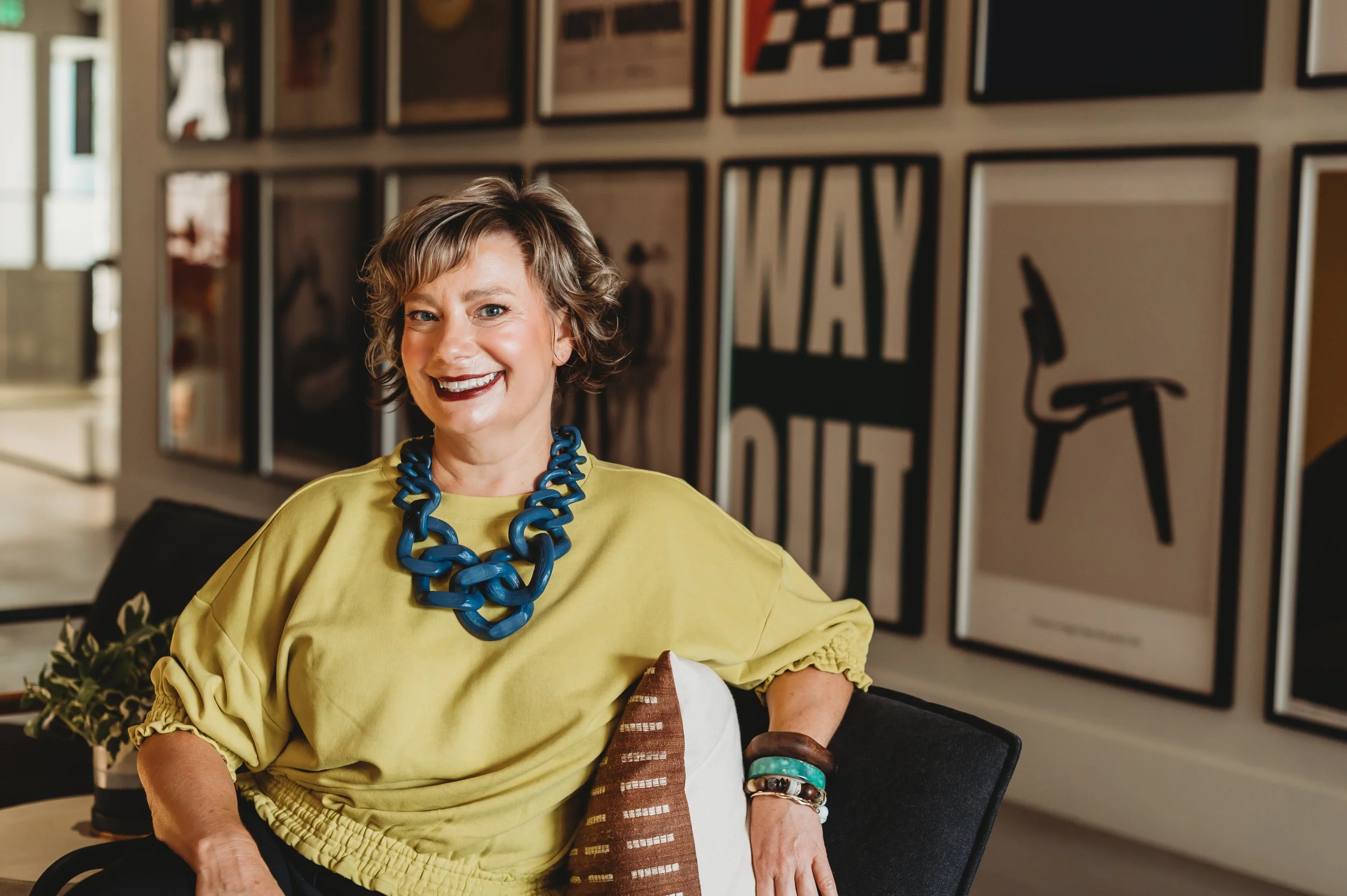 A woman with short curly hair wearing a yellow top and chunky blue necklace, smiling while sitting in a modern indoor space with framed artworks on the wall.