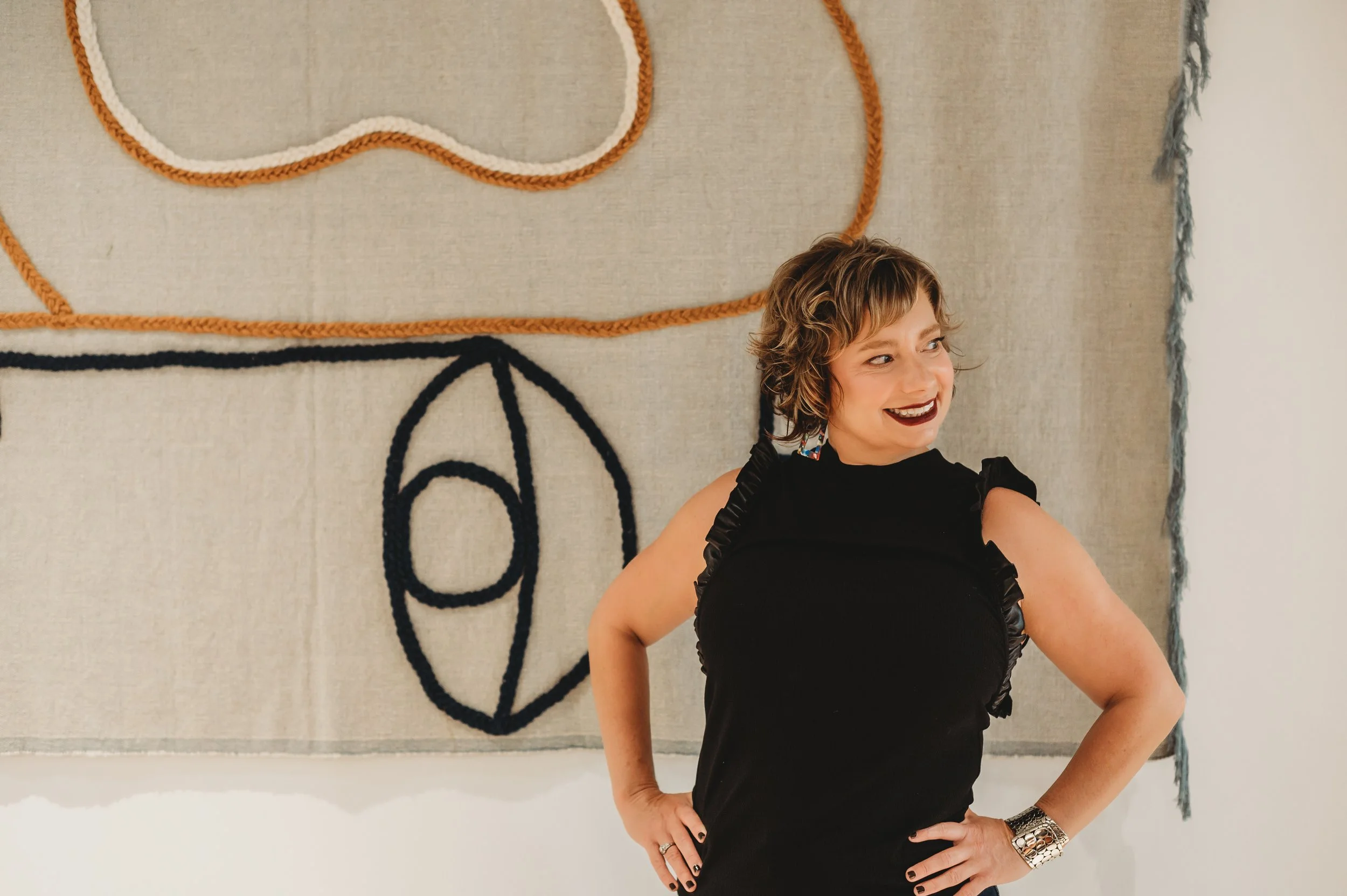 A woman with short, curly brown hair wearing a black dress and jewelry, smiling and standing with hands on hips in front of a textured wall with orange, white, and black rope art.