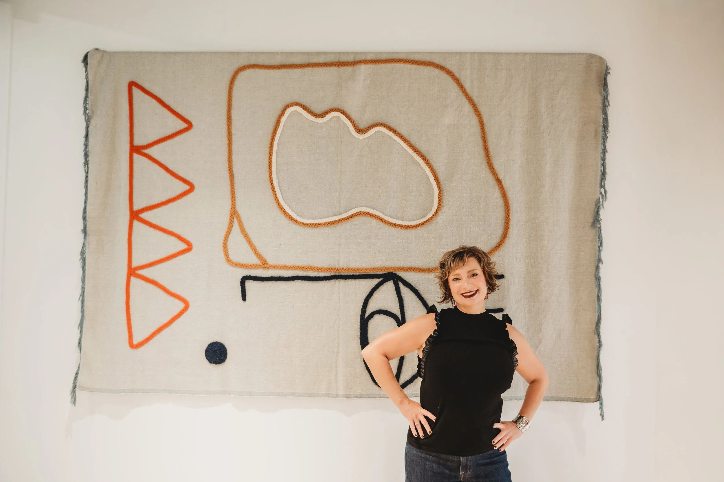 A smiling woman with short curly hair wearing a black sleeveless top posing in front of a wall hanging. The wall hanging is decorated with colorful, abstract embroidery designs on a beige background.