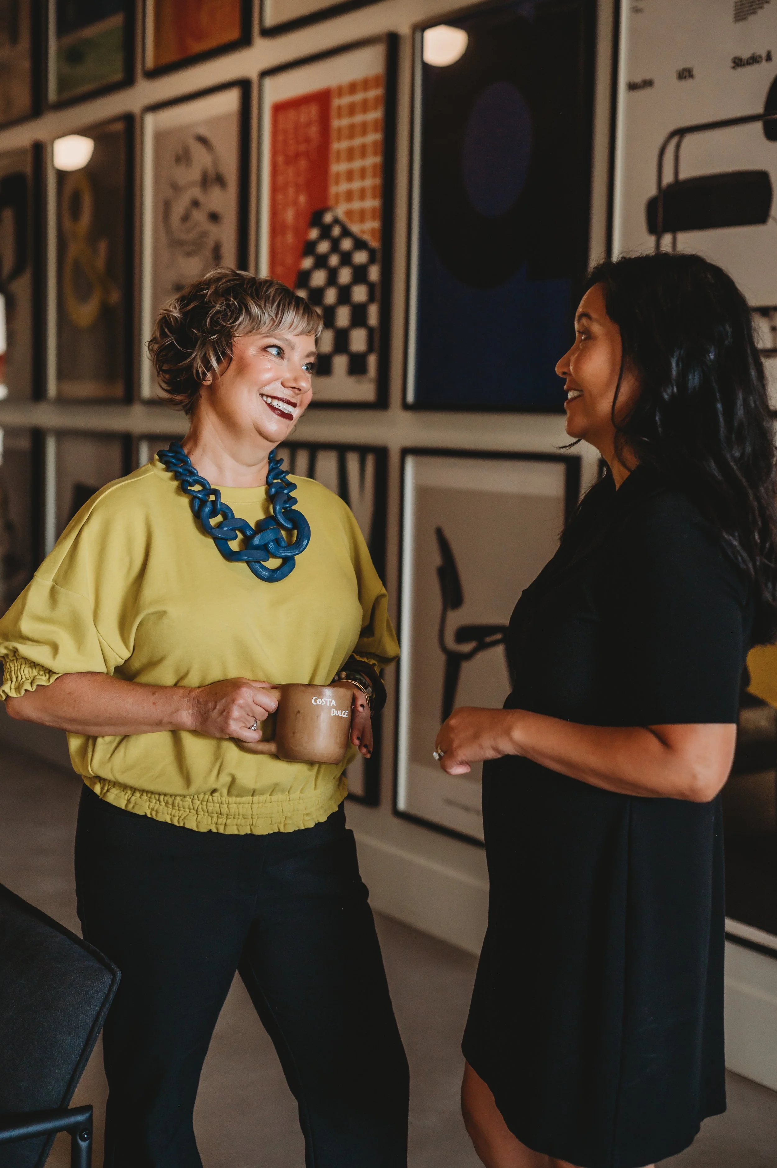 Two women engaged in conversation indoors, with one woman wearing a yellow top, a chunky blue necklace, and holding a coffee mug, and the other woman wearing a black dress, with a background of framed abstract artwork on the wall.