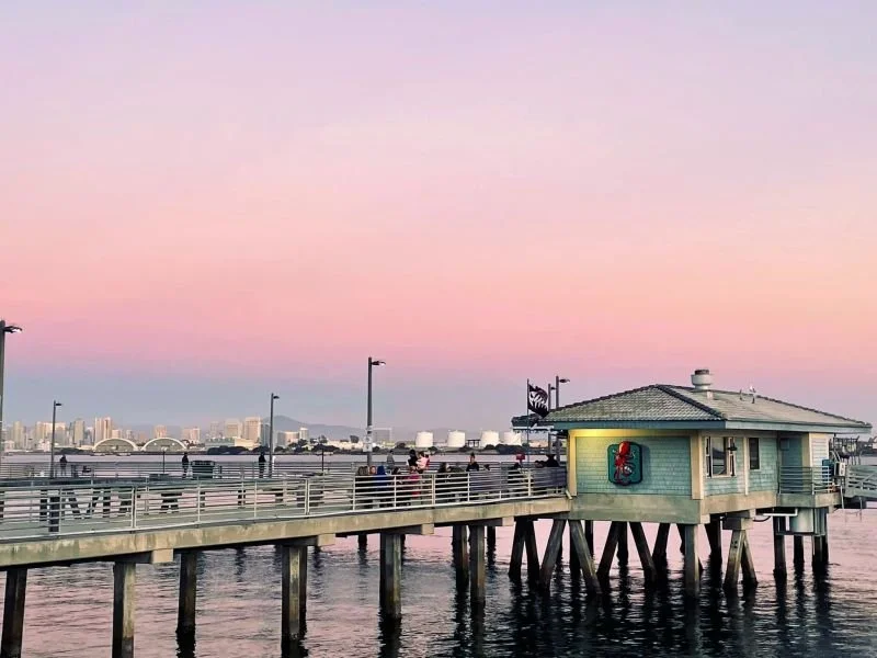 People standing on a pier at dusk with the city skyline in the background and a pink sky.