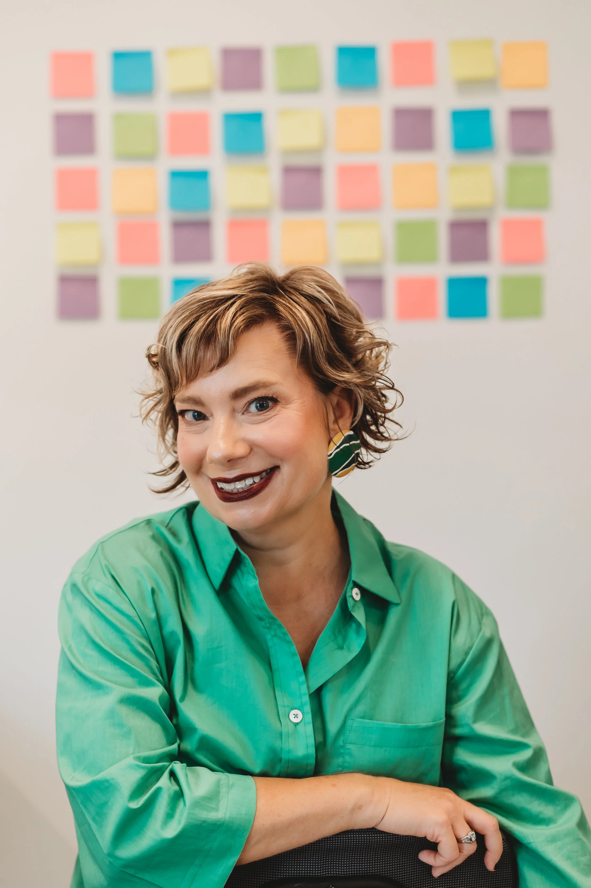 A woman with short, curly light brown hair and blue eyes, wearing a green shirt, sitting on a black chair, smiling, with a wall of colorful sticky notes behind her.