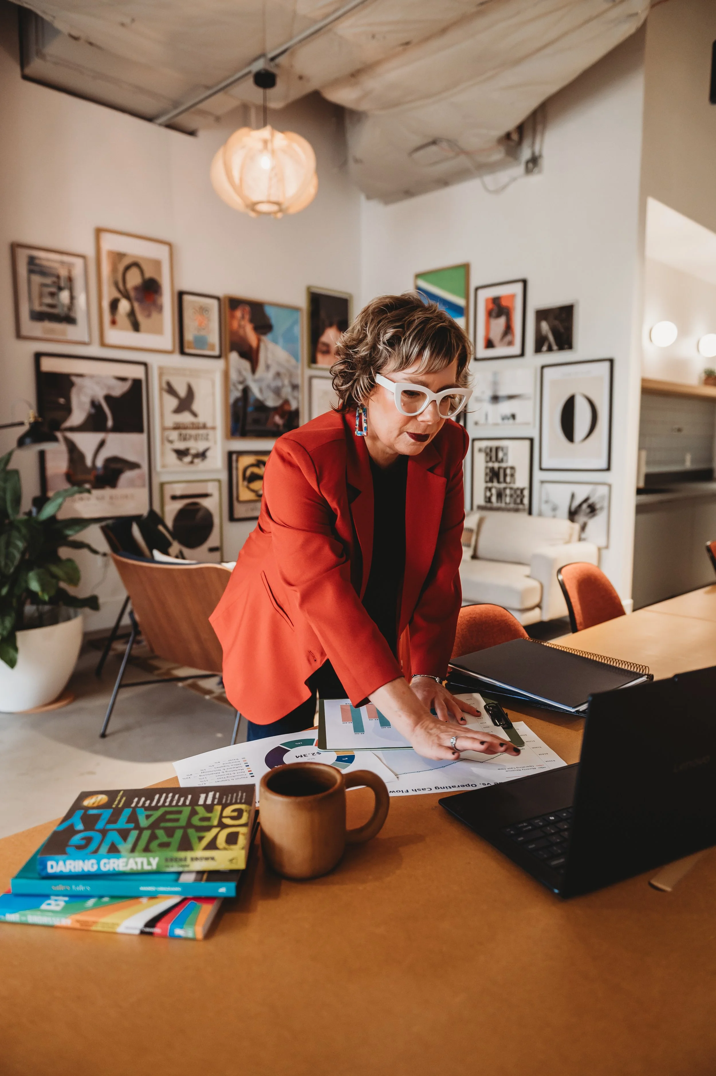A woman with curly hair and wearing glasses in a red blazer is working at a wooden table with charts, a laptop, books, and a coffee mug in an art-filled office or studio.