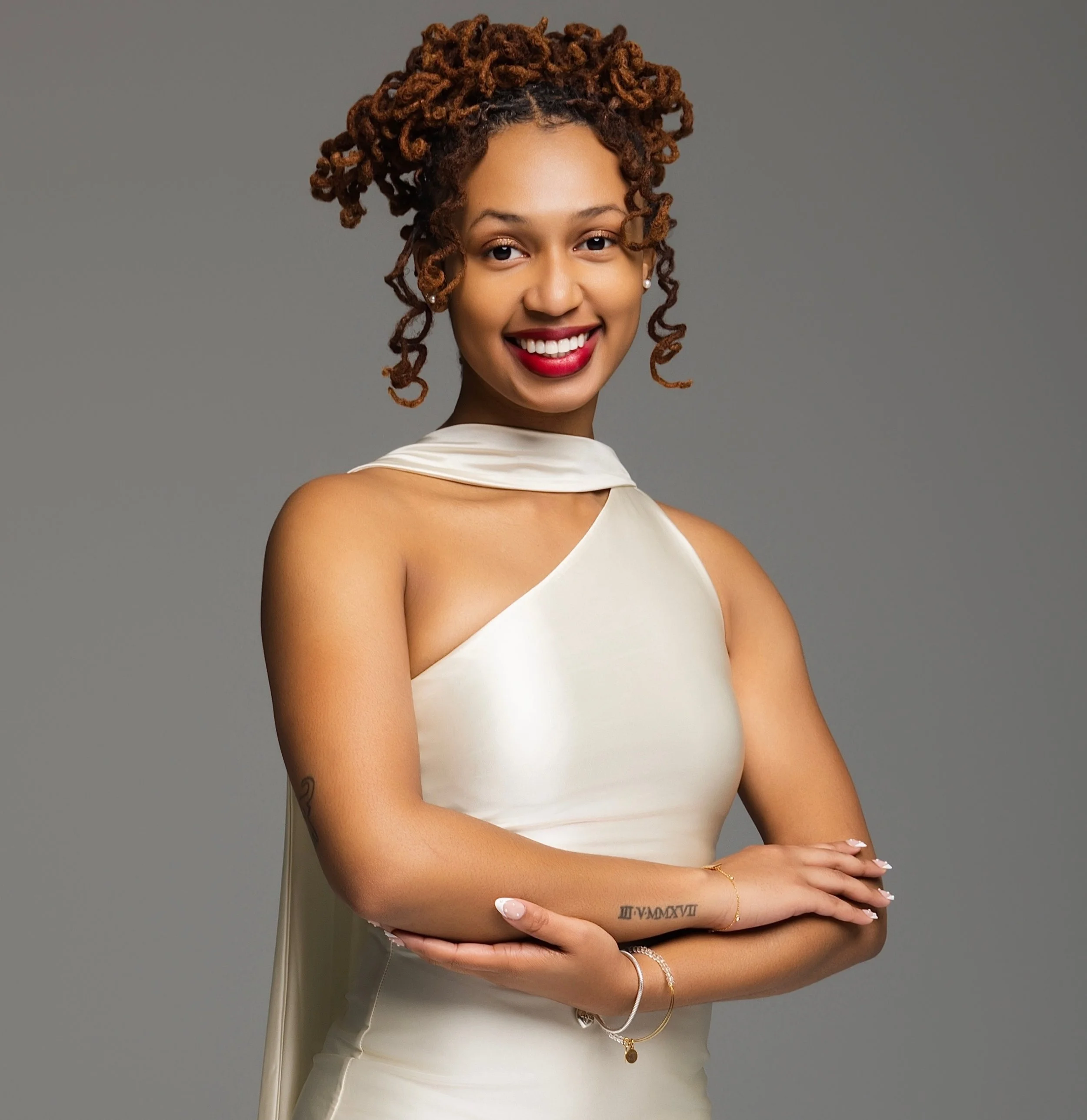 Close-up of a smiling woman with curly hair styled in an updo, wearing red lipstick, pearl earrings, a cream satin top, and gold jewelry, standing against a gray background.