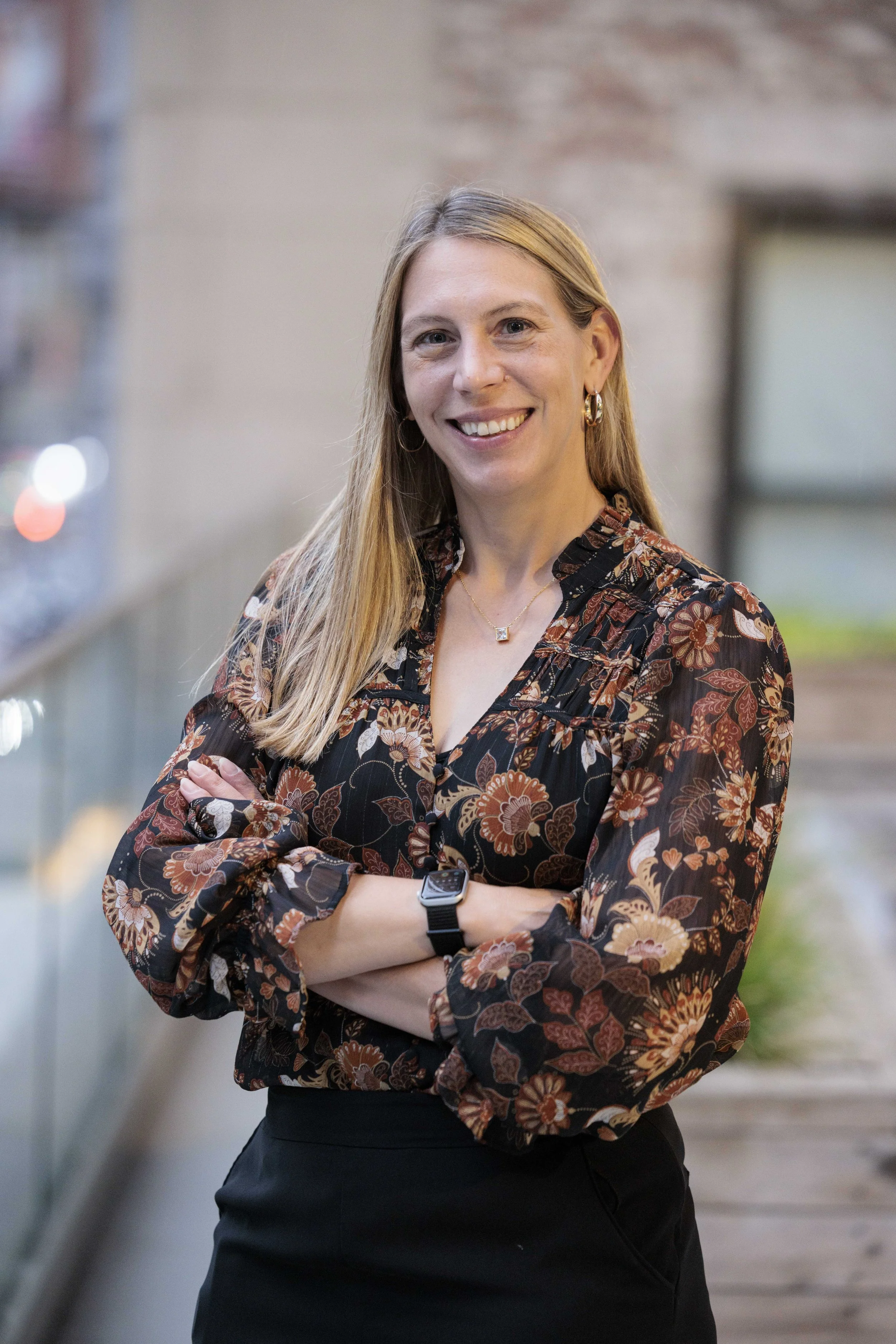 A woman with long blonde hair wearing a patterned shirt, black skirt, and smartwatch, standing outdoors with arms crossed and smiling.