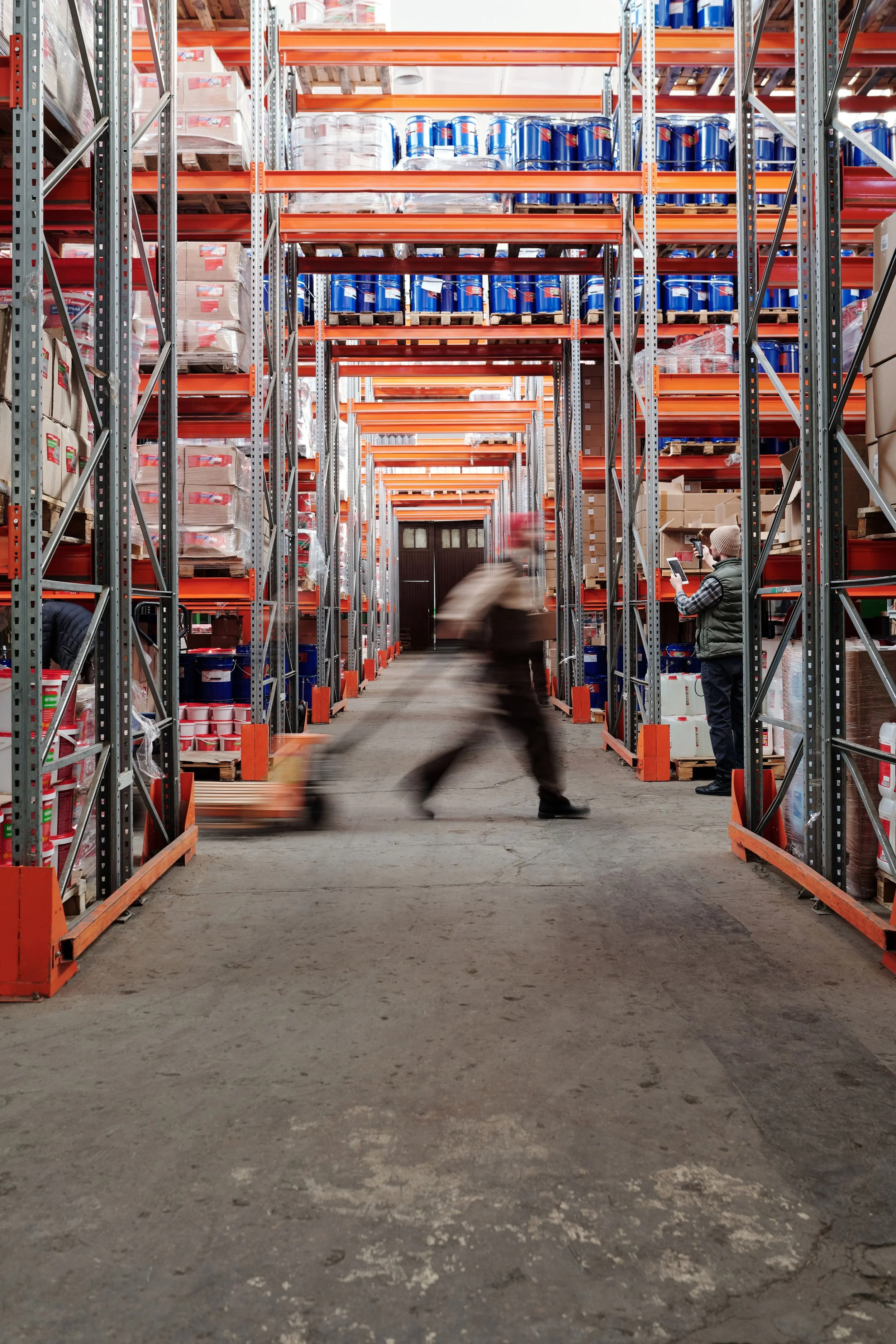 A large warehouse with tall orange and silver metal shelving filled with various boxes and containers. Several workers are present, one person is blurred indicating movement, and another is standing and looking at a smartphone.