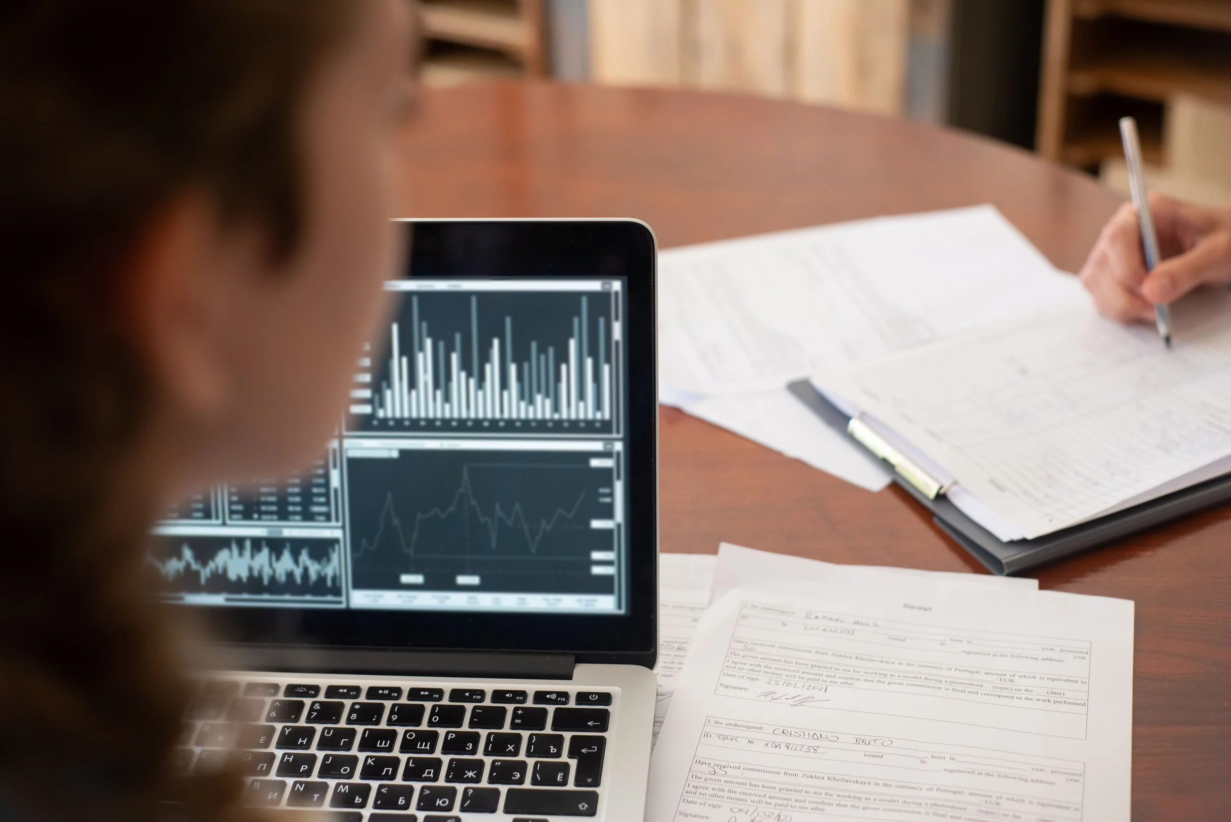 A person working at a desk with papers, a pen, and a laptop displaying graphs and charts.