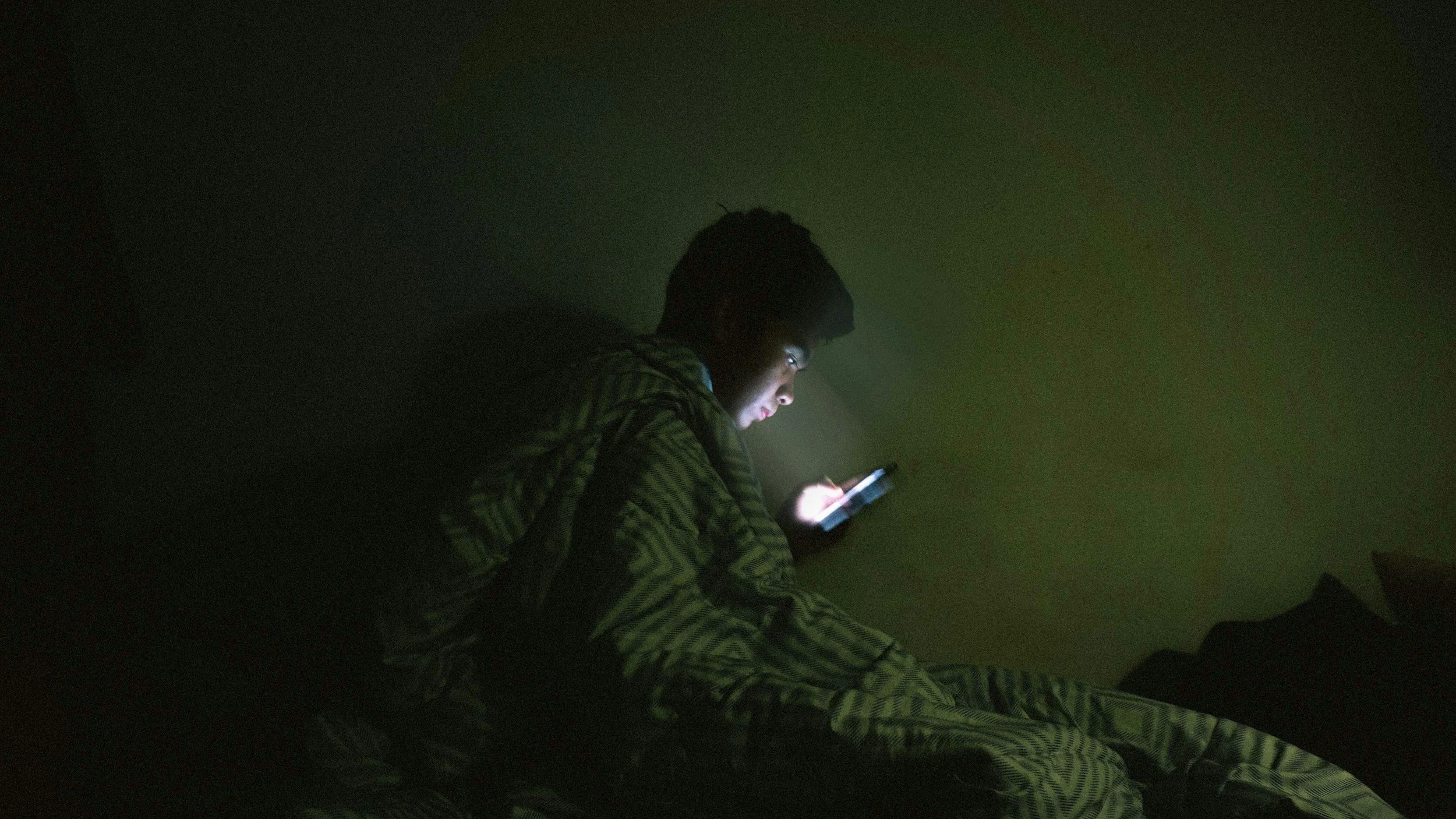 A person sitting on the floor in a dark room, illuminated only by the light of a smartphone they are holding.