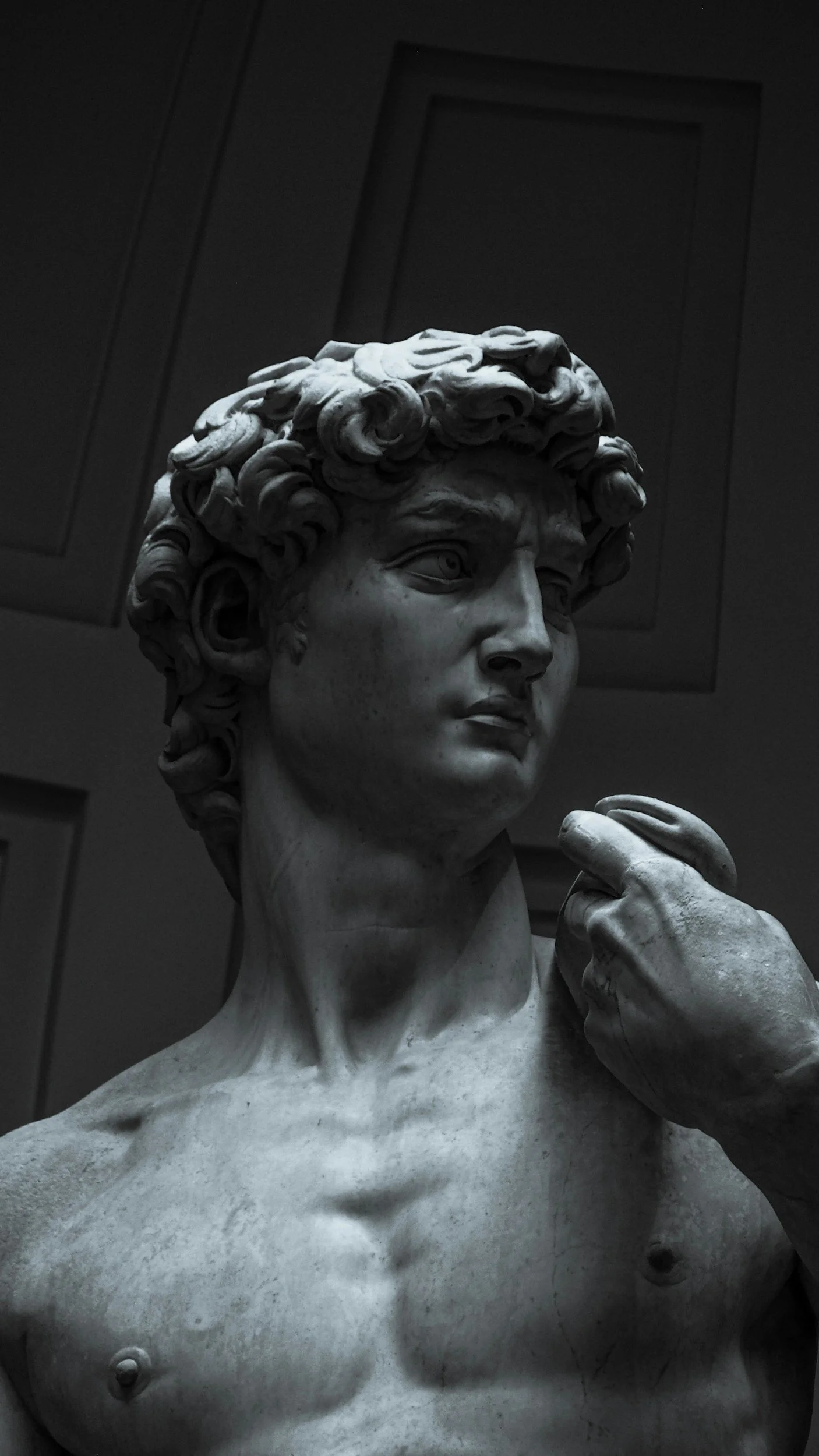 Close-up of a classical marble statue of a woman with curly hair, looking thoughtfully to the side, with her right hand near her face, set against a dark background.