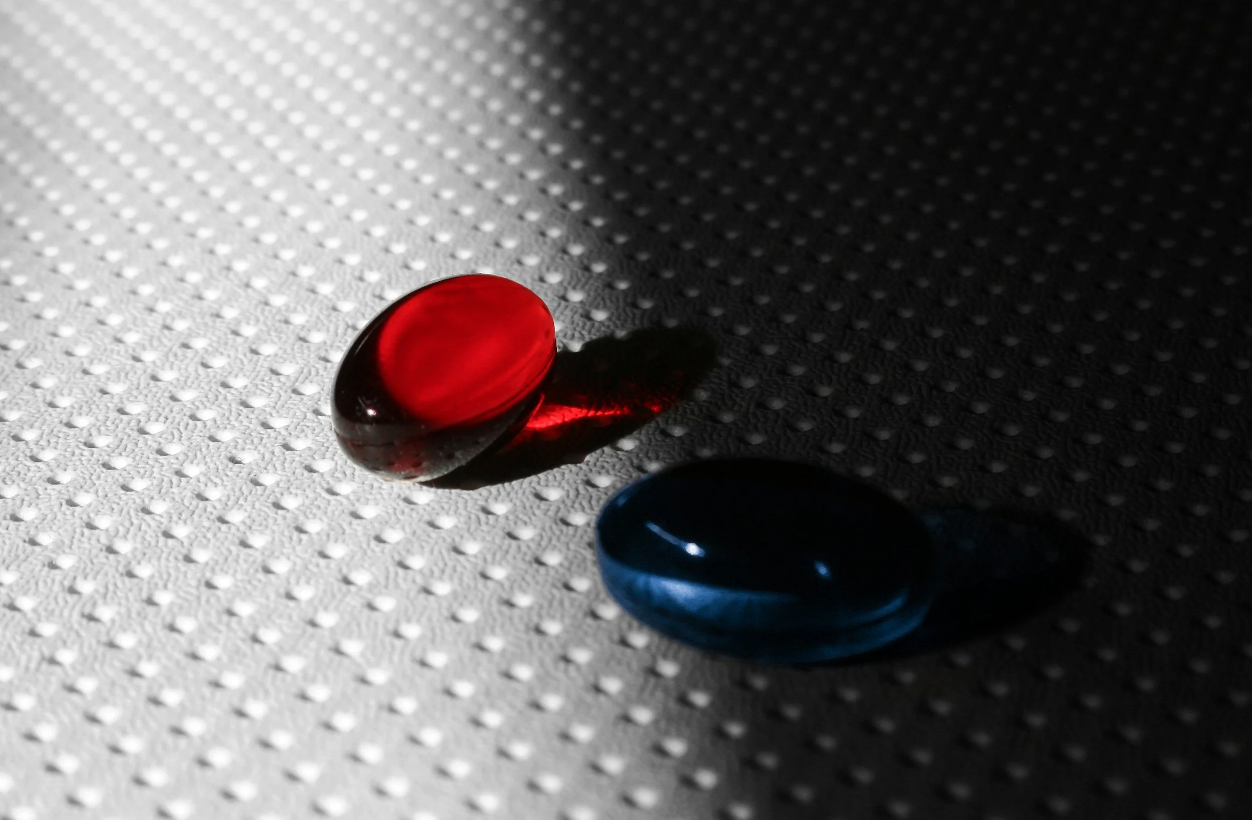 Close-up of a red glass marble on a textured white surface with a shadow, along with parts of a blue marble and a dark sphere.