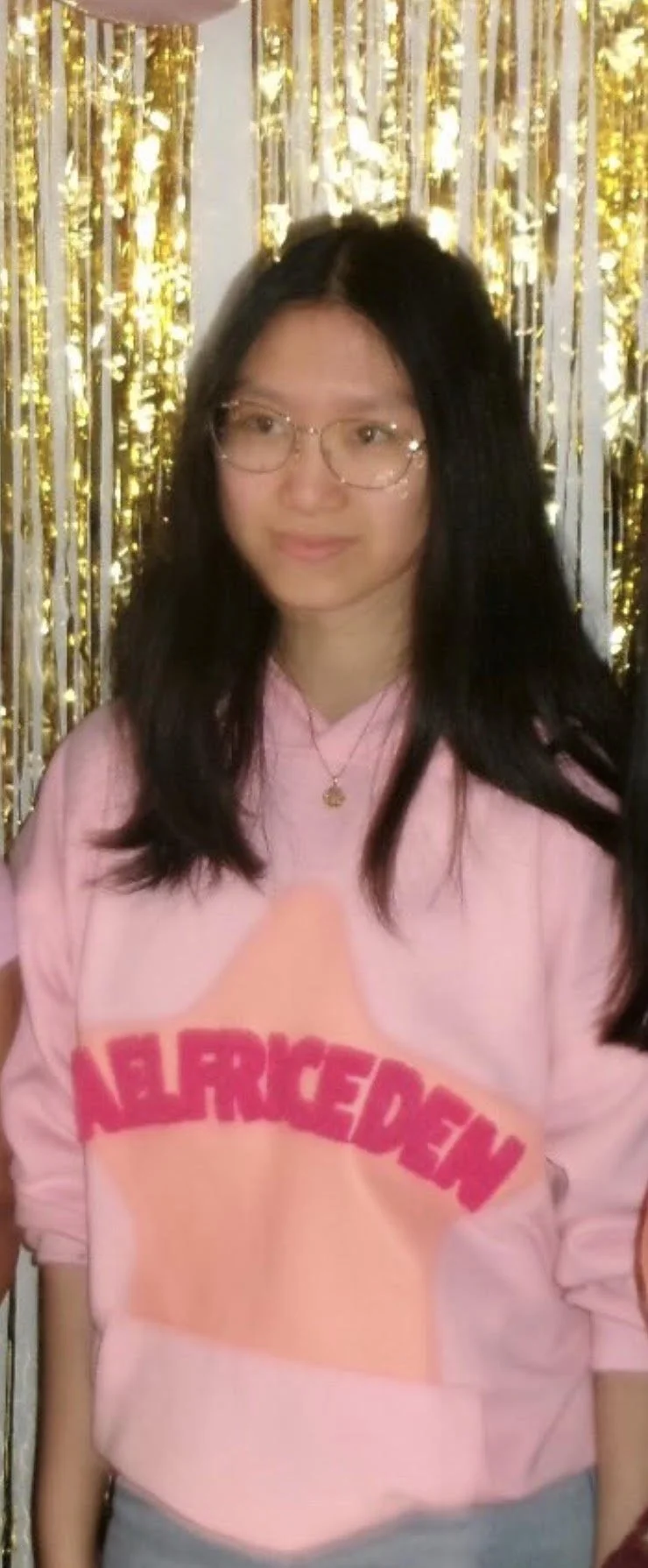 Young woman with long dark hair and glasses, wearing a pink Abercrombie hoodie and a gold necklace, standing in front of a shiny gold and silver curtain backdrop.