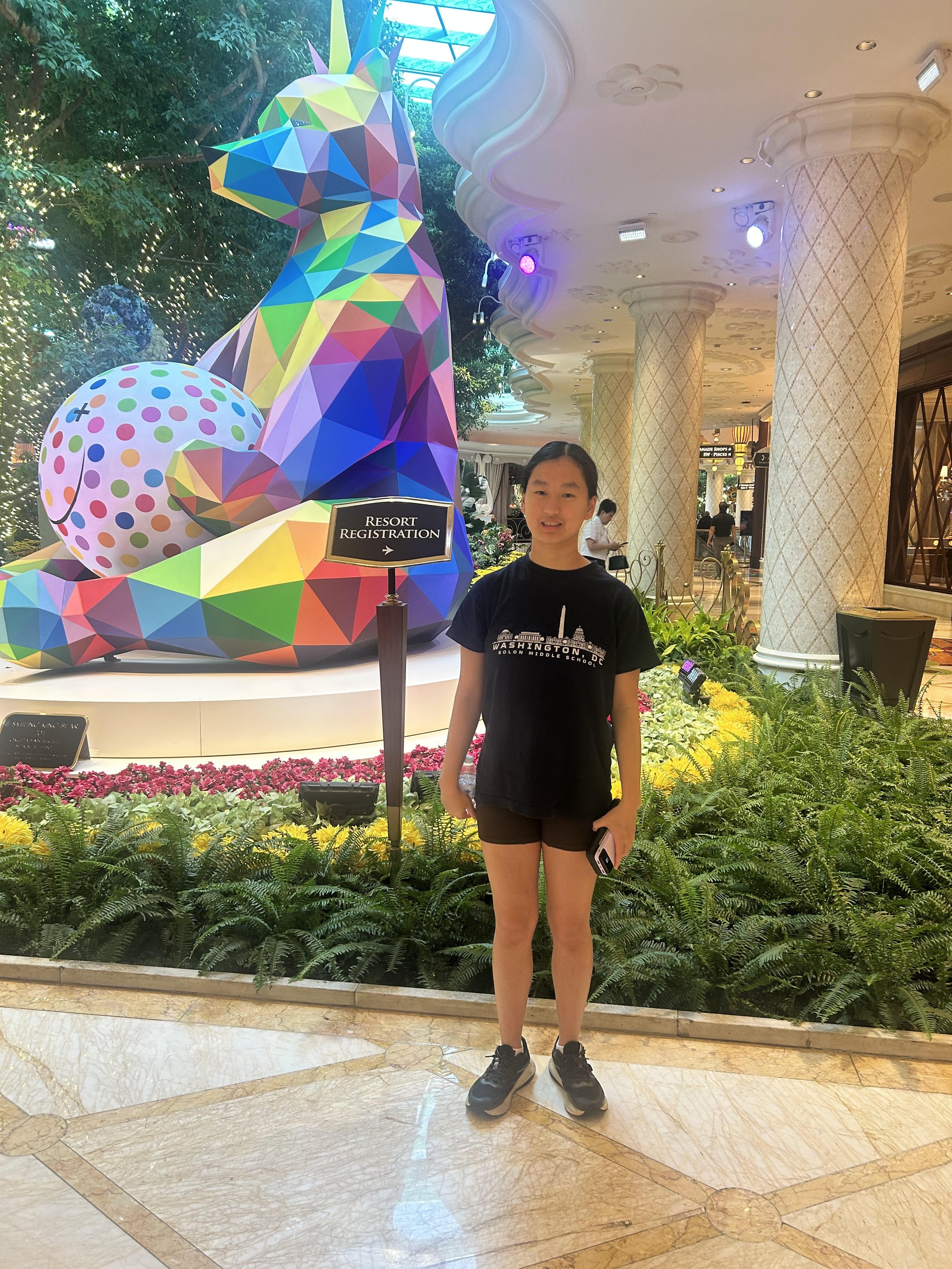 A young girl is standing inside a luxurious hotel or casino lobby in front of a colorful geometric unicorn sculpture and a sign that says 'Resort Registration.' She is wearing a black T-shirt with a cityscape graphic and shorts, holding a phone, with a backdrop of decorative columns, plants, and a polished marble floor.
